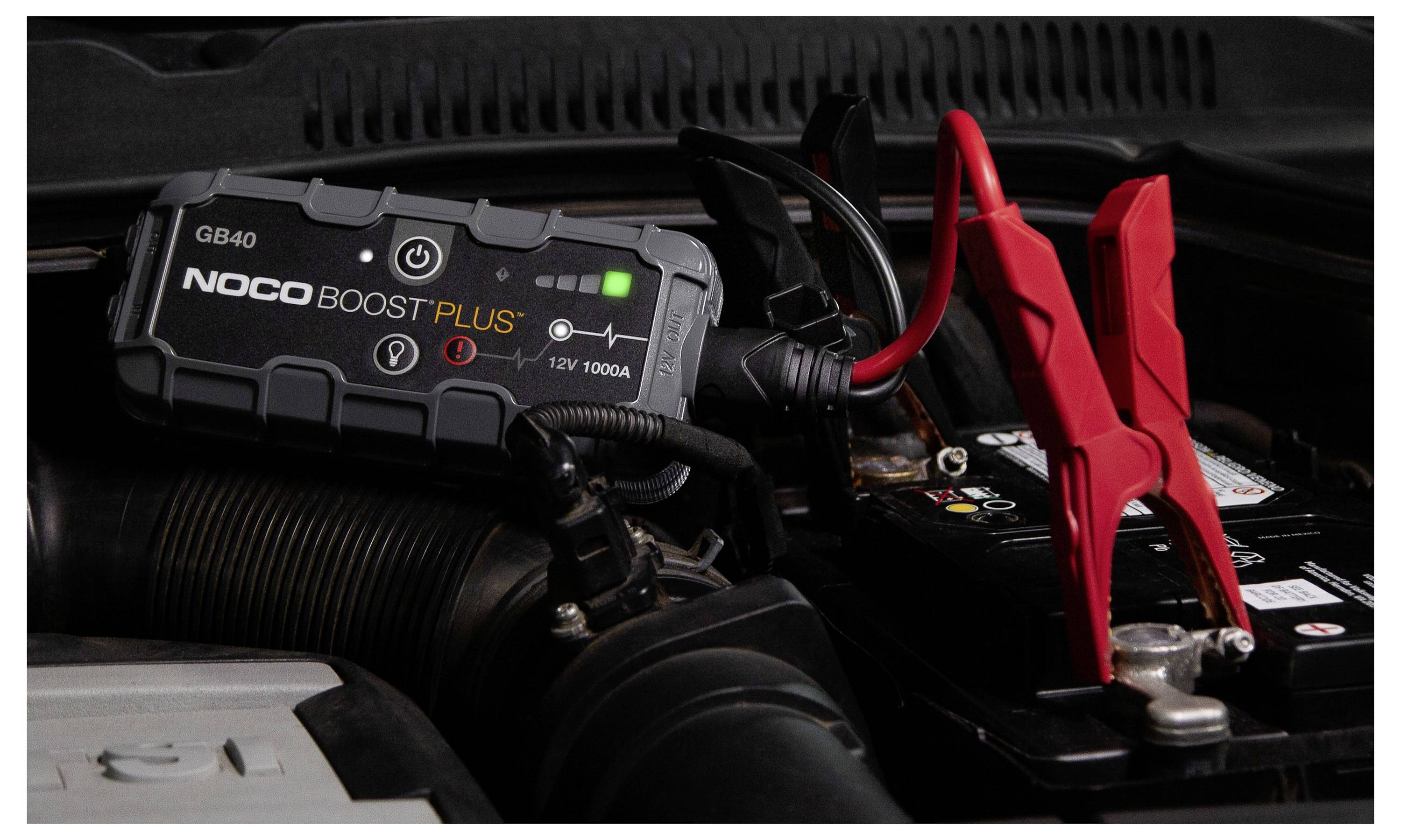 Ein NOCO Boost Plus Starthilfegerät ist mit roten und schwarzen Klemmen an einer Autobatterie befestigt und zeigt den Motorraum des Fahrzeugs.