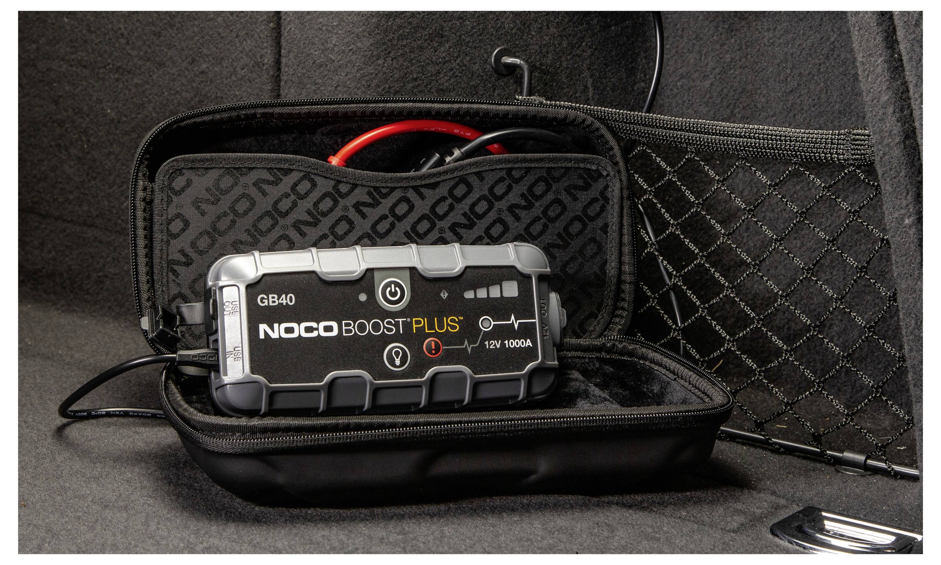 Ein NOCO Boost Plus GB40 Starthilfegerät in einer schwarzen Tasche mit Reißverschluss mit Kabeln und Klemmen, auf einer dunklen Oberfläche aufgeklappt dargestellt.