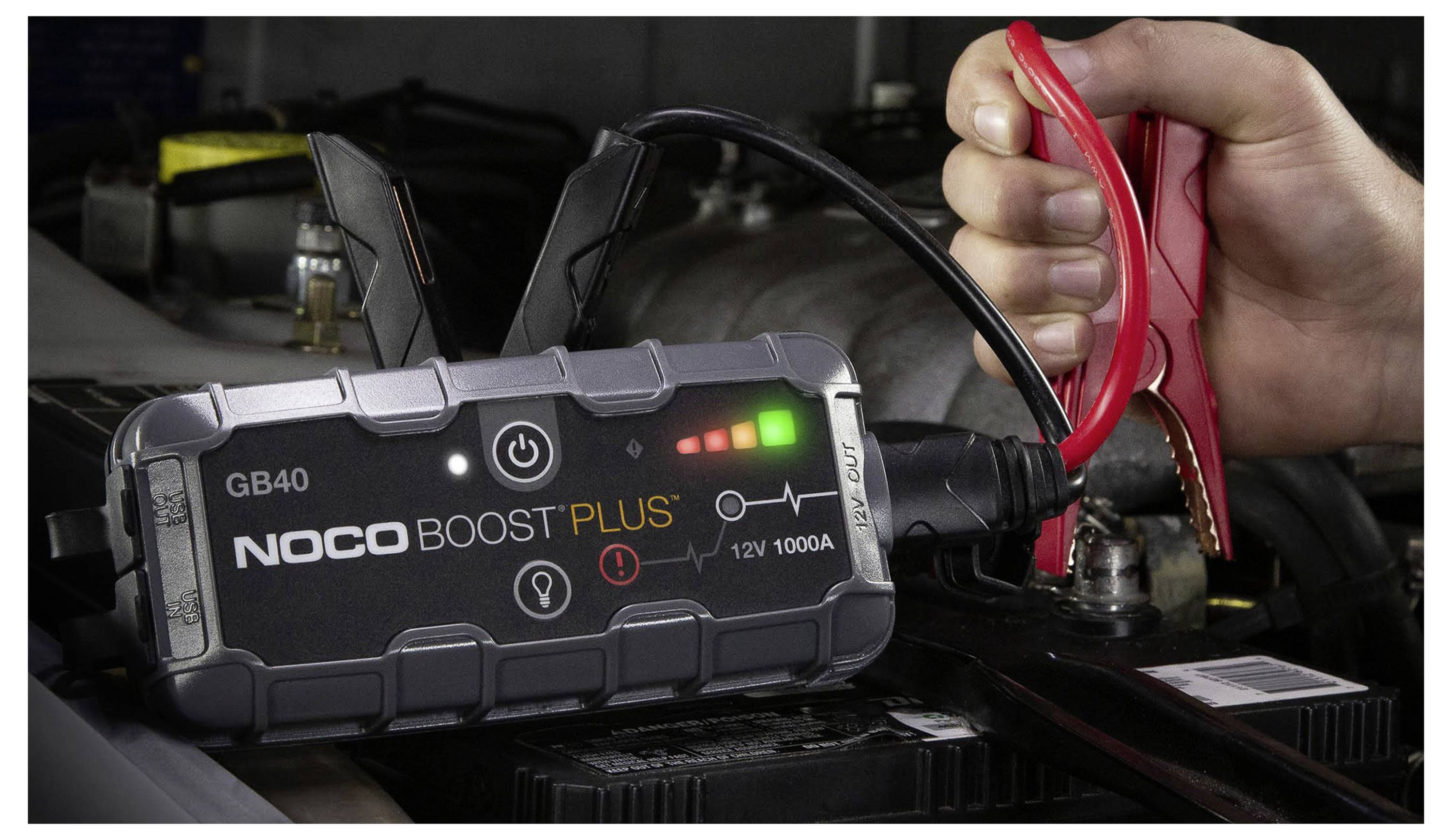 Eine Hand verbindet rote Überbrückungskabel an eine Autobatterie mit einem NOCO Boost Plus GB40 tragbaren Starthilfegerät in einem Fahrzeugmotorraum.