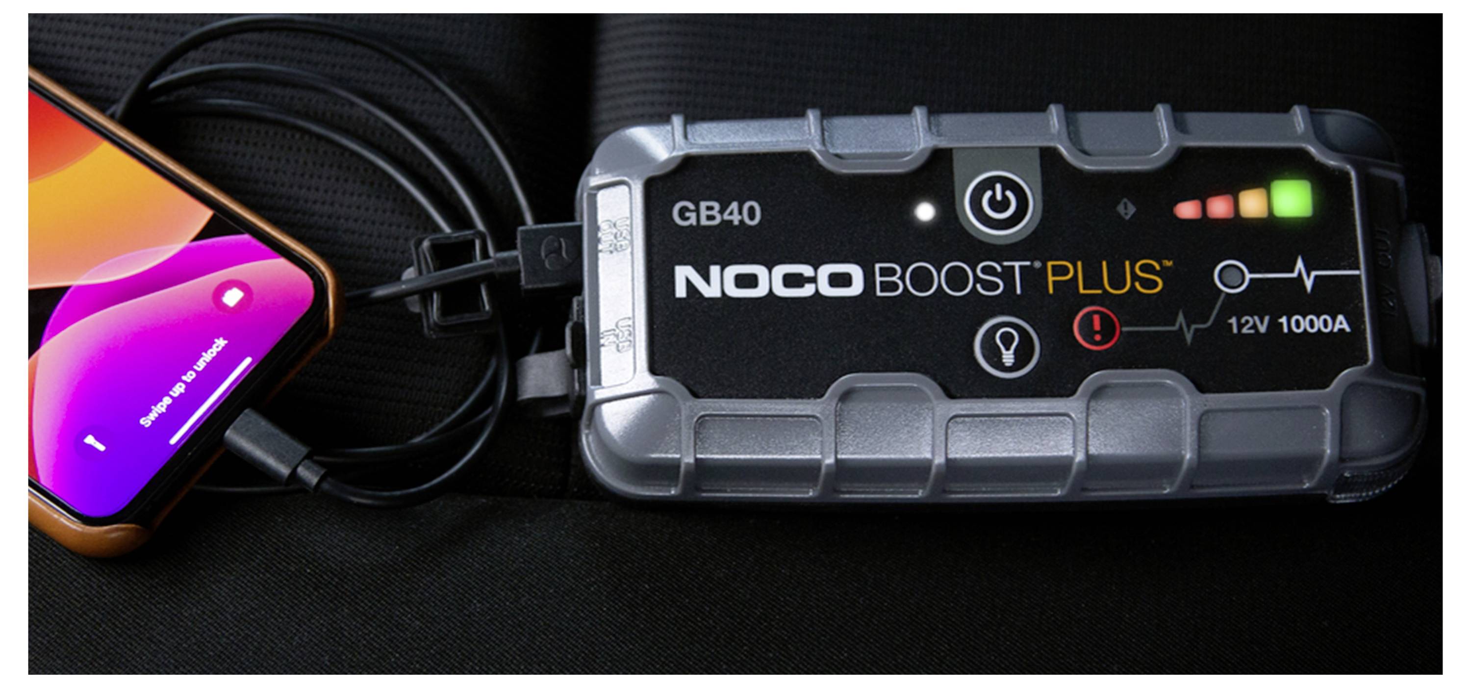 Ein tragbarer NOCO Boost Plus GB40 Starthilfegerät, das mit einem Kabel mit einem Smartphone verbunden ist, mit leuchtenden Stromanzeigeleuchten.
