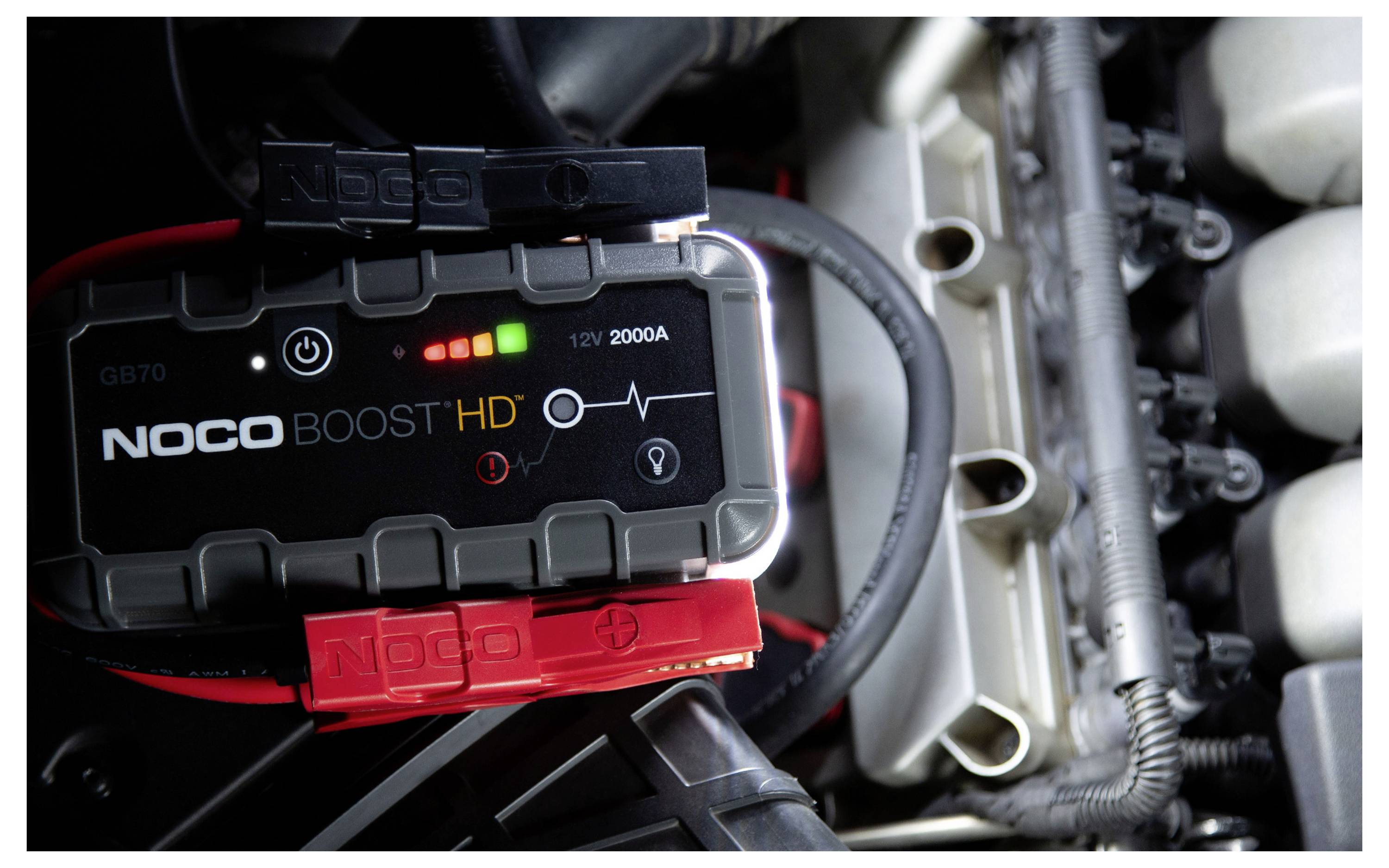 Ein NOCO Boost HD Starthilfegerät, das in einem Motorraum angeschlossen ist, zeigt Batterieladungsanzeigen und Klemmkabel, die bereit sind, das Fahrzeug zu starten.