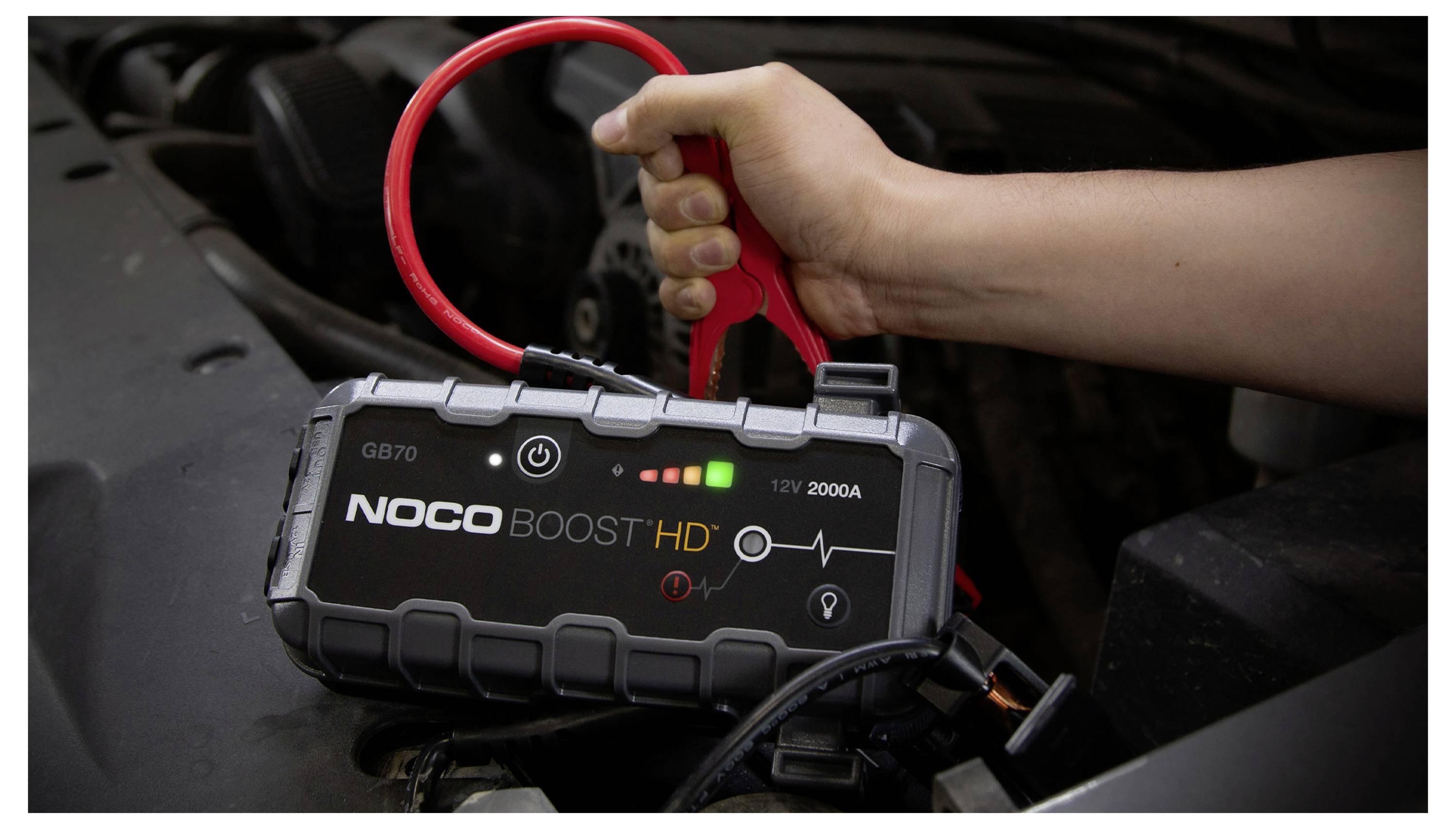 Hand, die einen NOCO Boost HD tragbaren Starthilfe hält, der mit einer Autobatterie verbunden ist, mit Ladeanzeigen und 12V 2000A Kennzeichnung.
