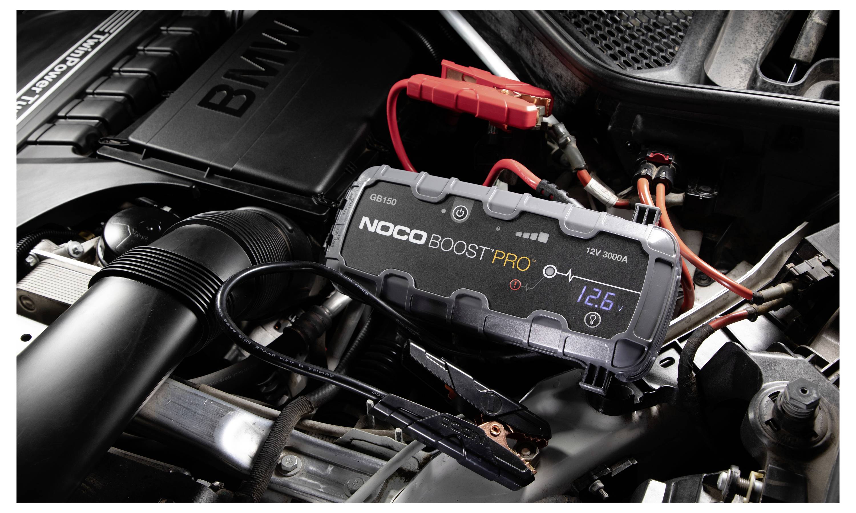 Ein NOCO Boost Pro Starthilfegerät ist mit roten und schwarzen Klemmen an einer Autobatterie im Motorraum angeschlossen und zeigt die Batteriespannung auf seinem Display an.