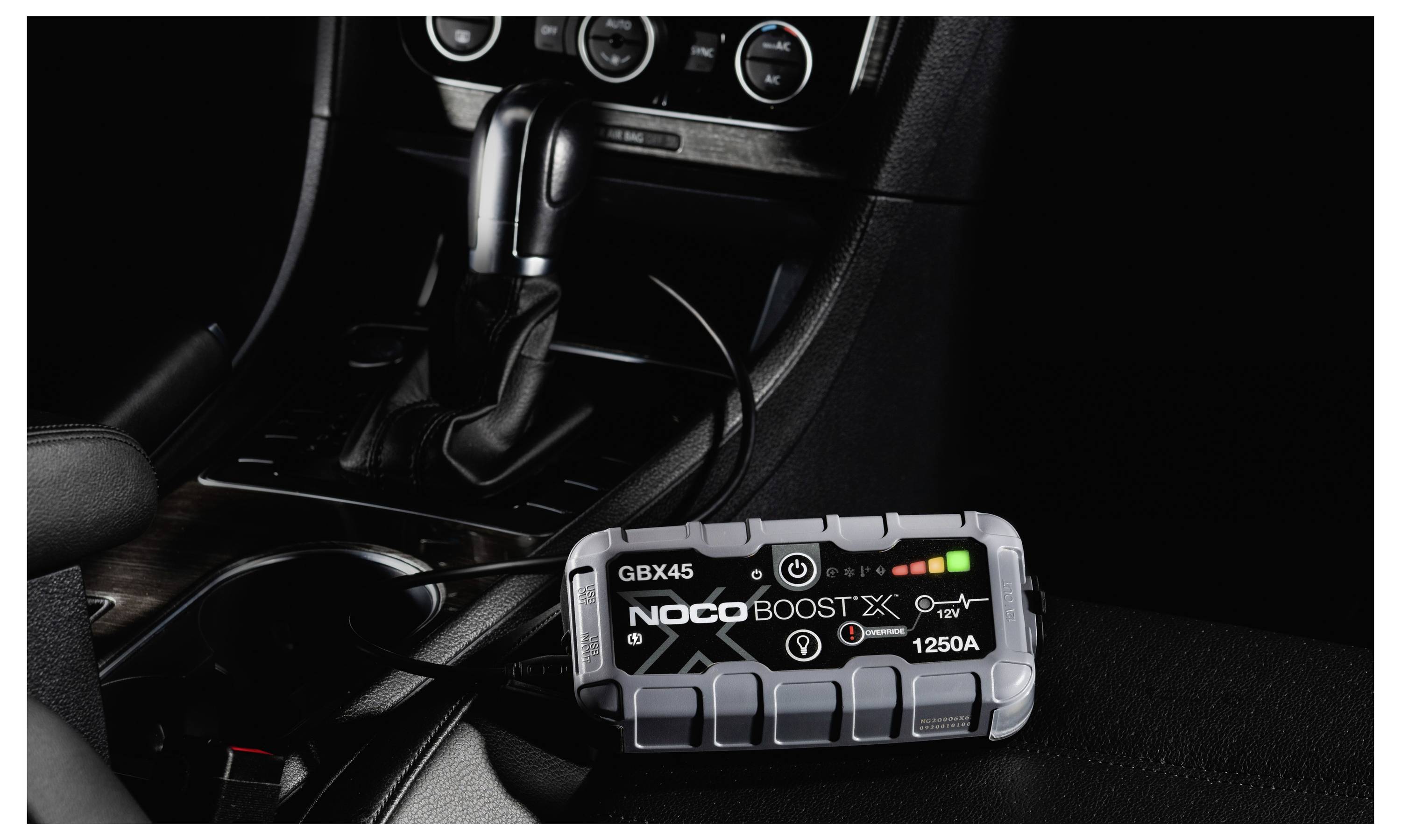 Tragbarer NOCO Boost X 1250A Starthilfegerät, das auf einem Autositz neben der Mittelkonsole und dem Schalthebel ruht, mit angeschlossenem Kabel.