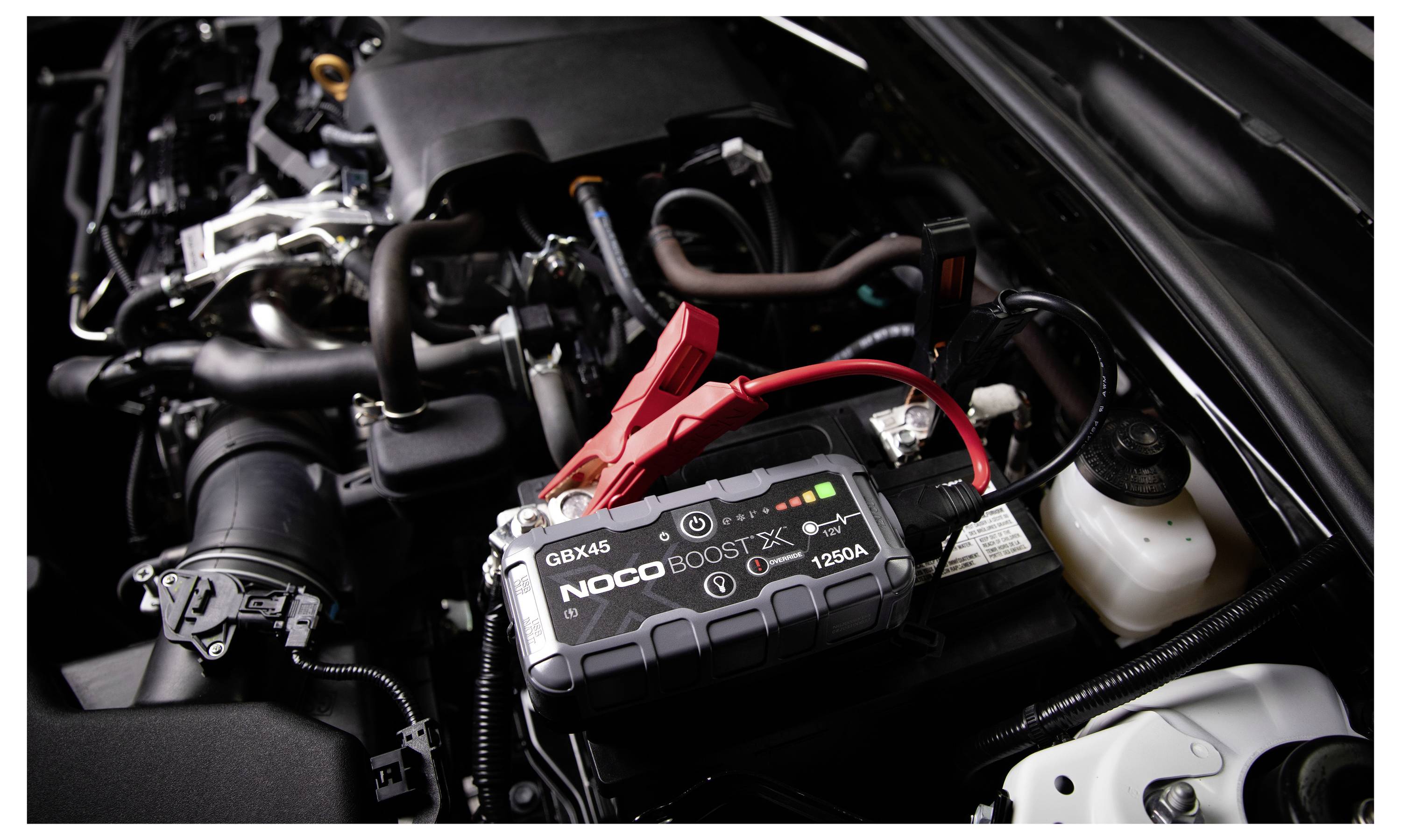 Ein tragbarer NOCO Boost X Autobatterie-Starthilfegerät mit roten und schwarzen Klemmen ist im Motorraum eines Fahrzeugs angeschlossen.