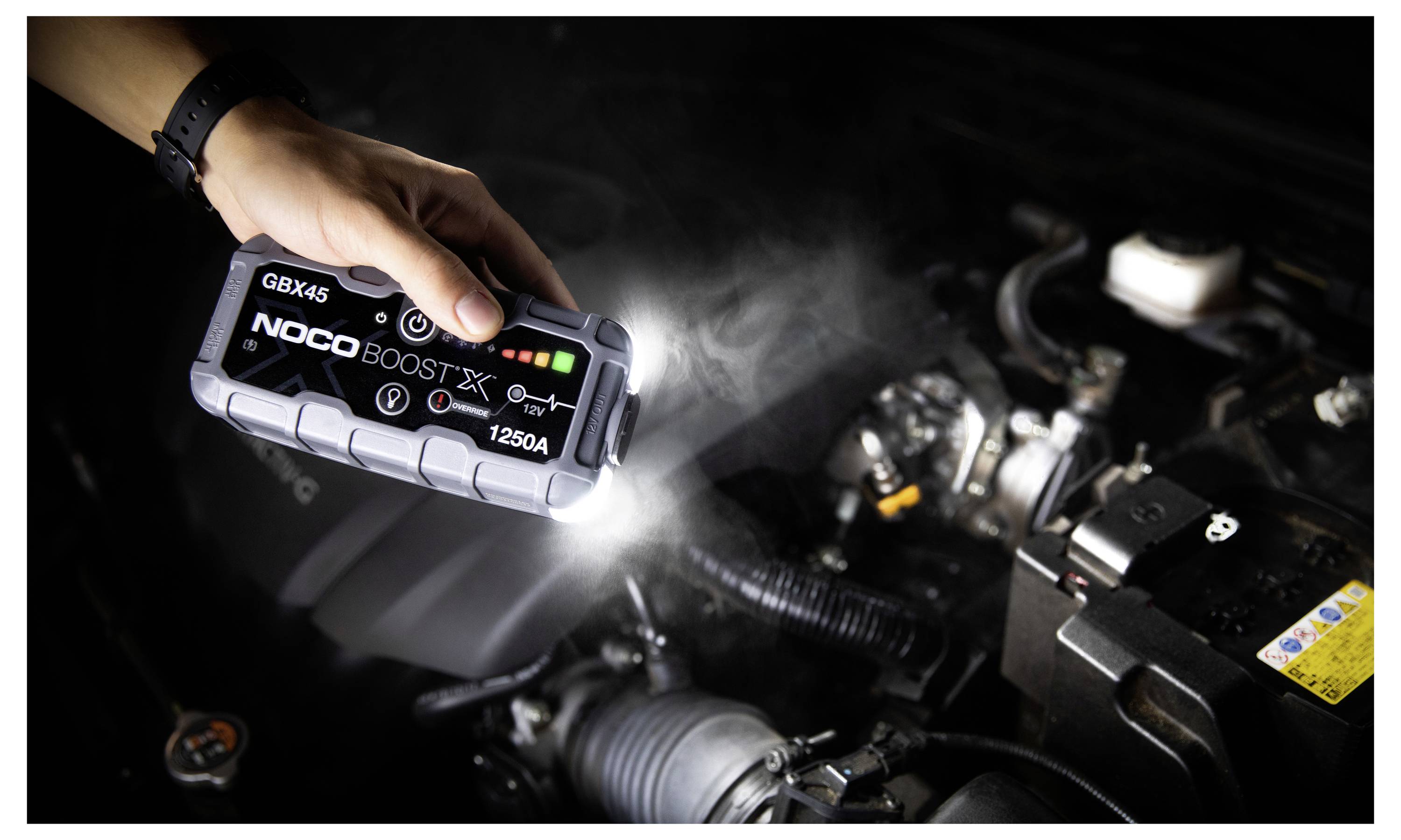 Hand hält einen NOCO Boost X GBX45 Starthilfegerät in einem Motorraum eines Autos, wobei dessen Taschenlampe zur Inspektion von Komponenten verwendet wird.