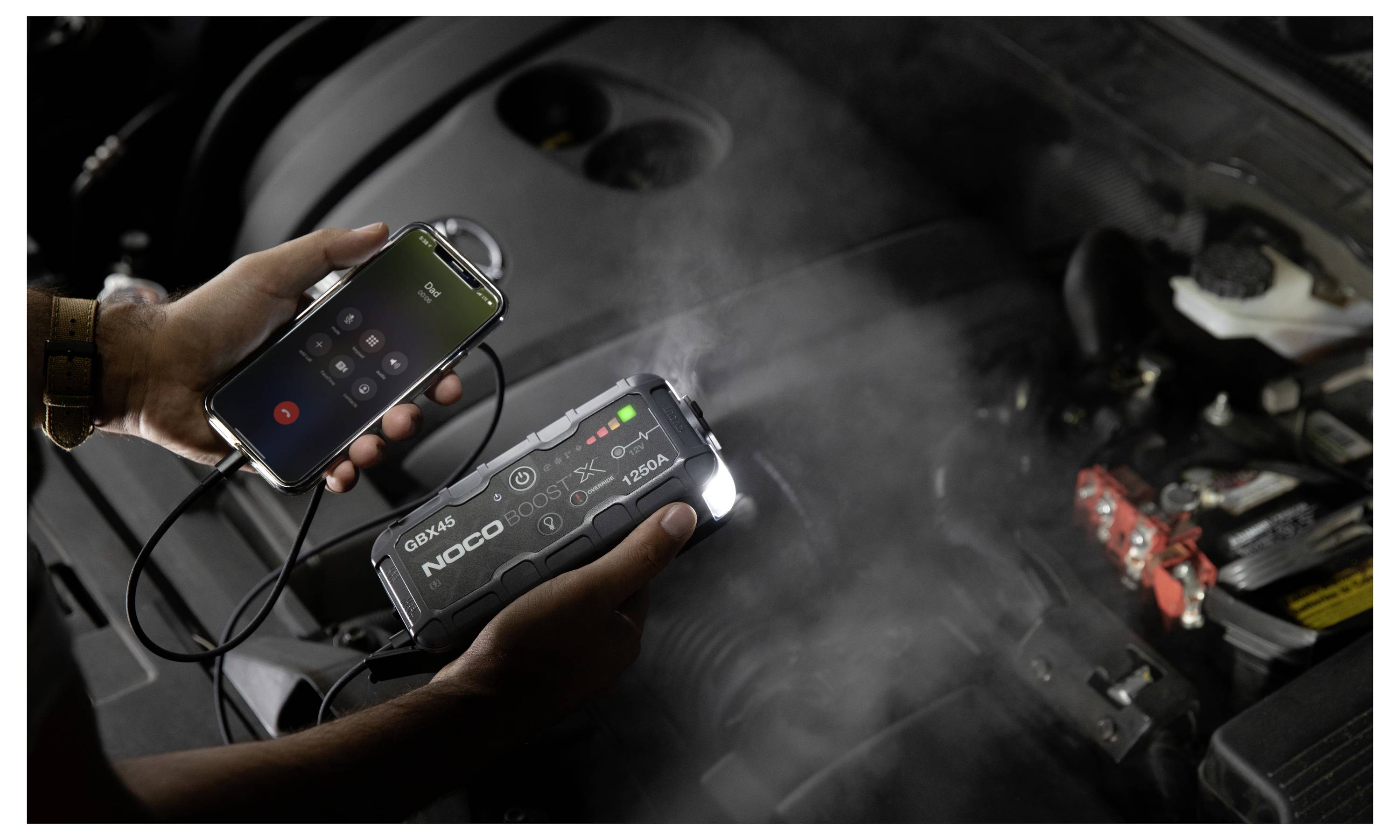 Person nutzt ein NOCO Starthilfegerät und eine Smartphone-App in der Nähe einer Autobatterie unter der Motorhaube, wobei ein digitales Display den Ladestatus anzeigt.