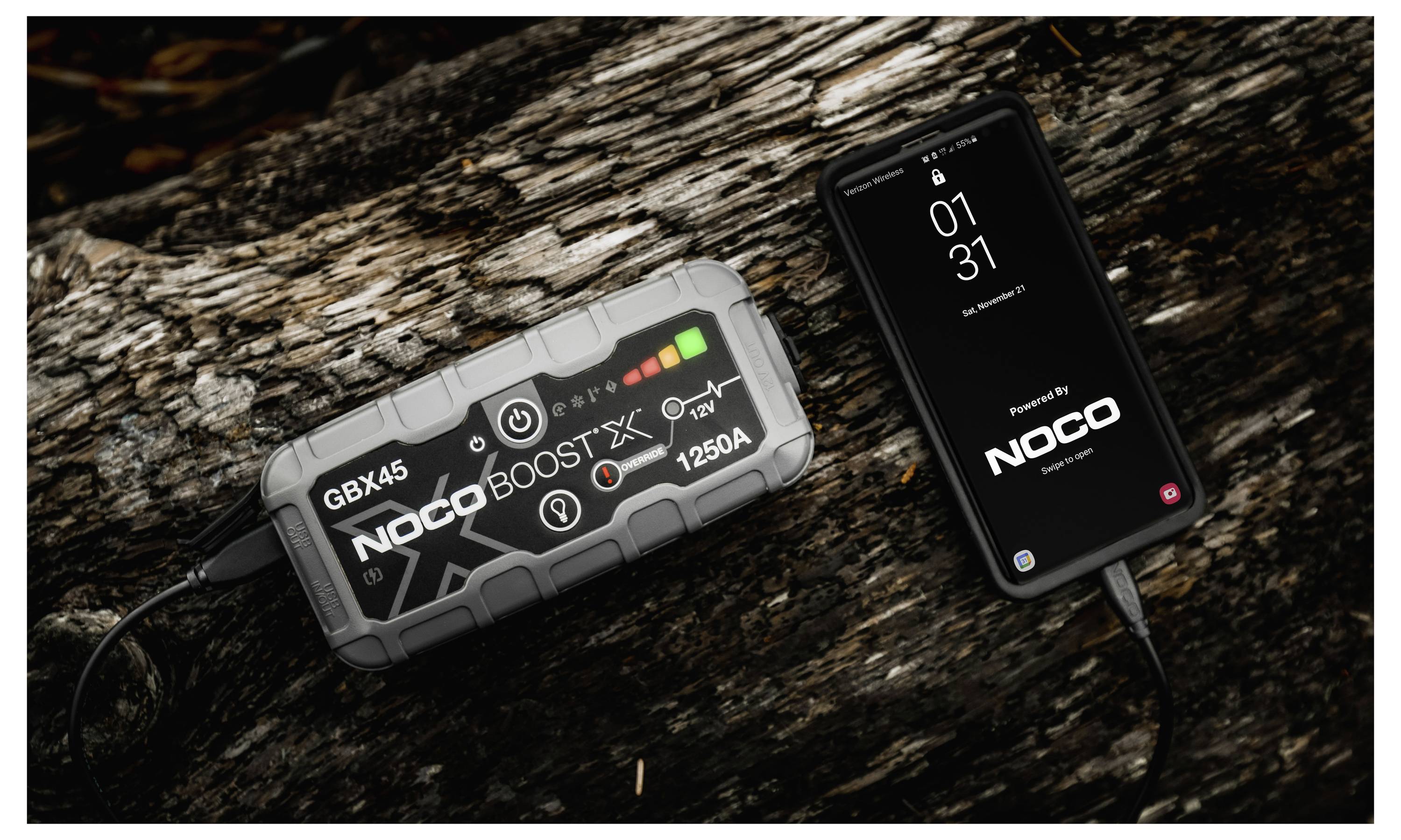 NOCO GBX45 Starthilfe mit einem Smartphone auf einem rauen Baumstamm verbunden, mit Lade-/Statussymbolen, die die Stromübertragung anzeigen.