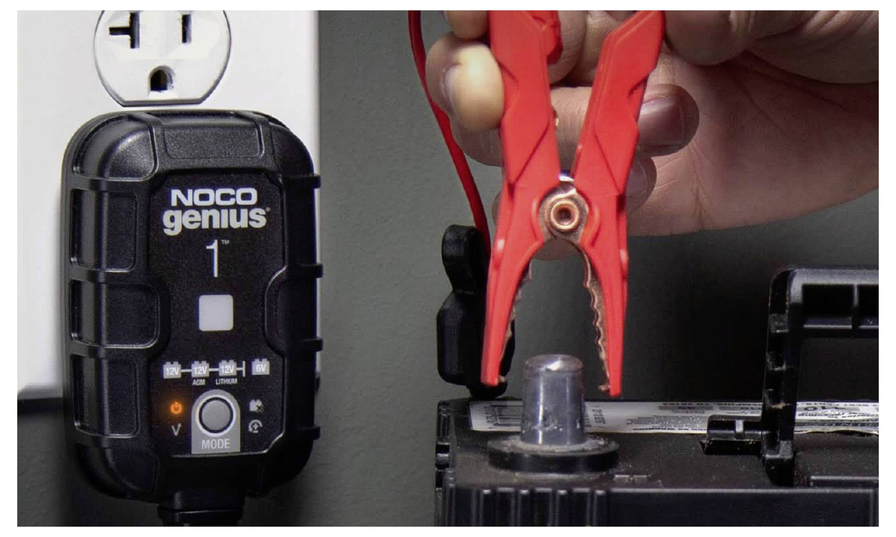 Hand hält rote Starthilfeklemme in der Nähe einer Autobatterie, mit einem NOCO Genius 1 Batterieladegerät, das links in die Steckdose eingesteckt ist.