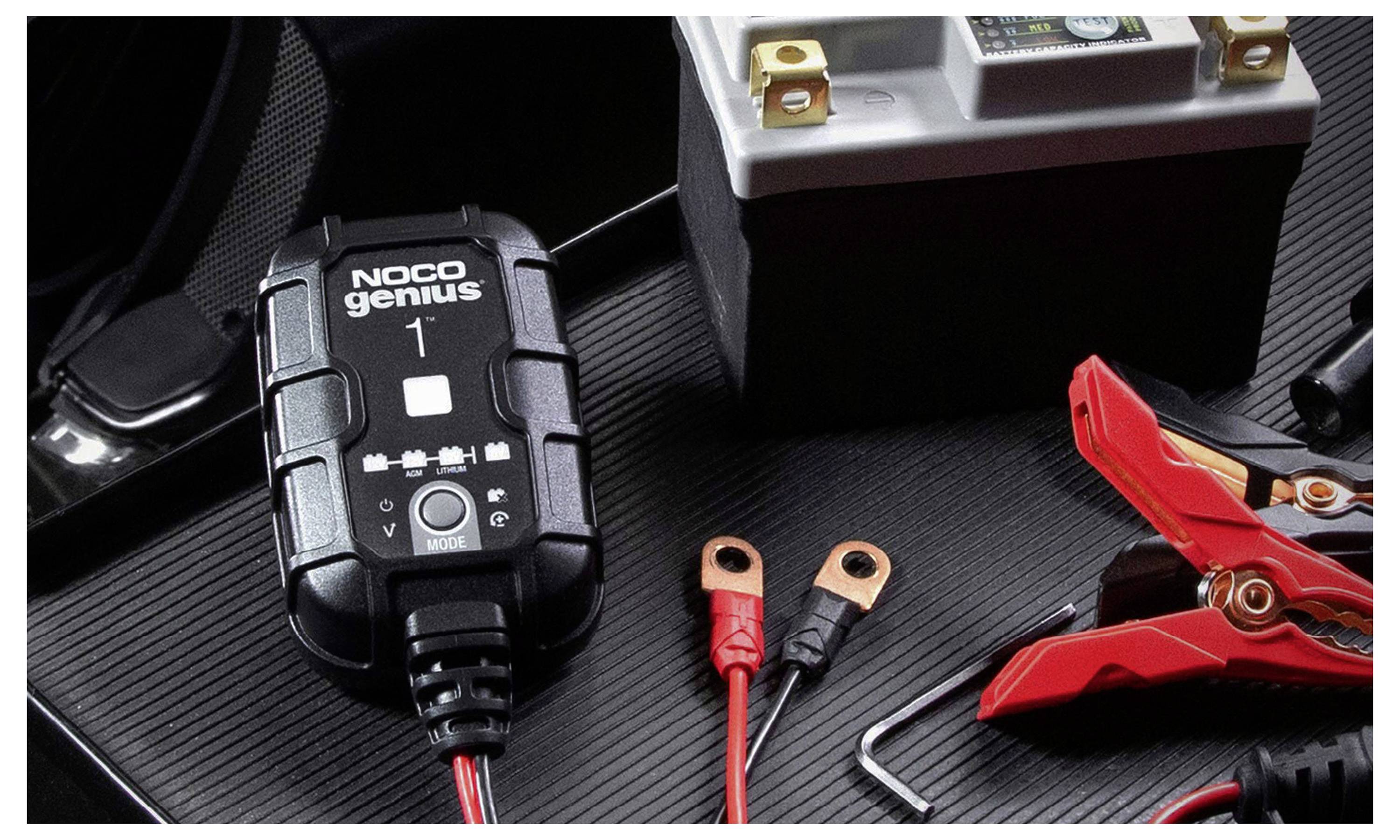 Ein NOCO Genius 1 Batterieladegerät mit roten und schwarzen Ringklemmen und einer roten Klemme auf einer strukturierten schwarzen Oberfläche.