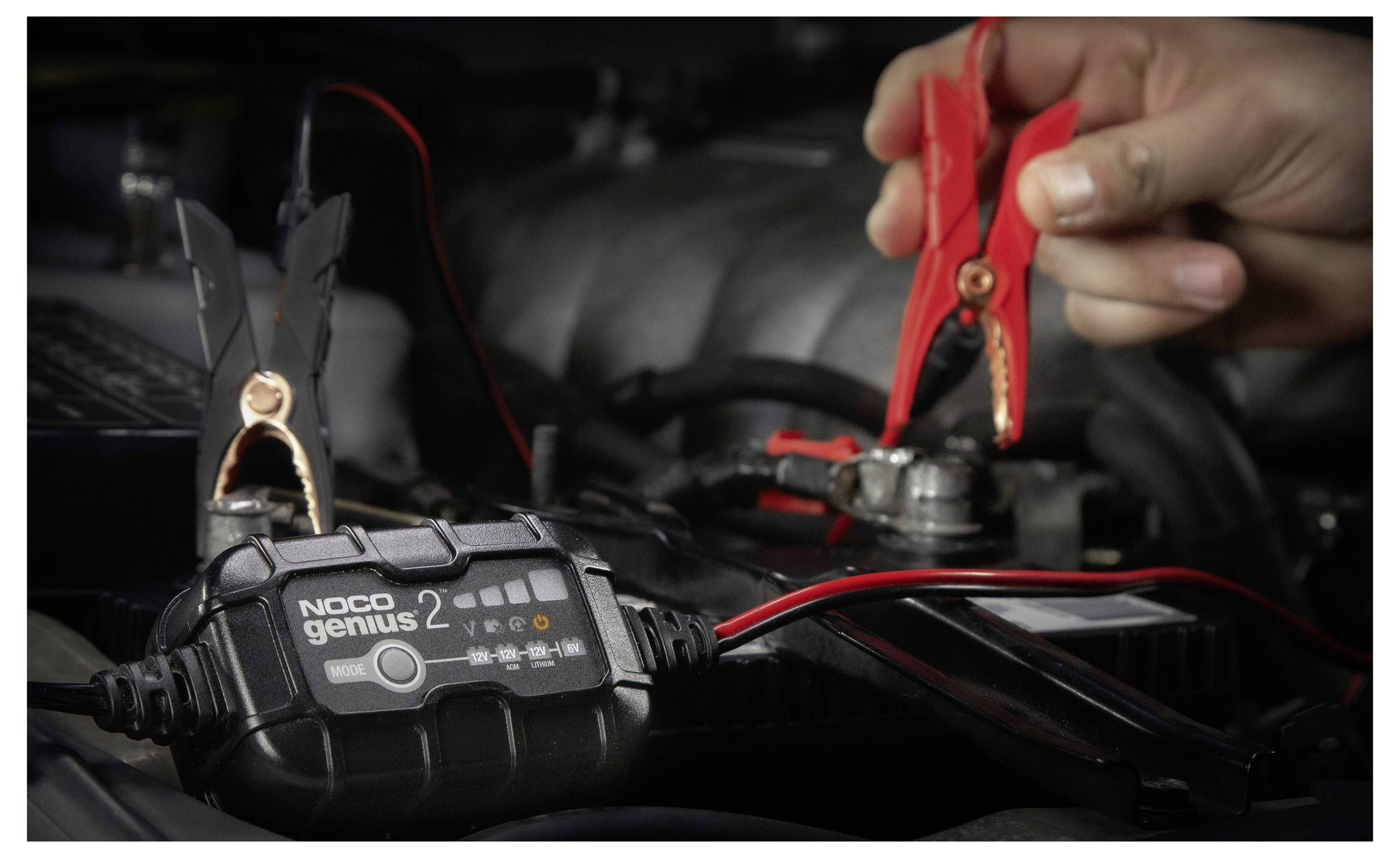 Hände, die rote und schwarze Klemmen verwenden, um ein NOCO Genius Batterieladegerät an eine Autobatterie in einem Motorraum anzuschließen.