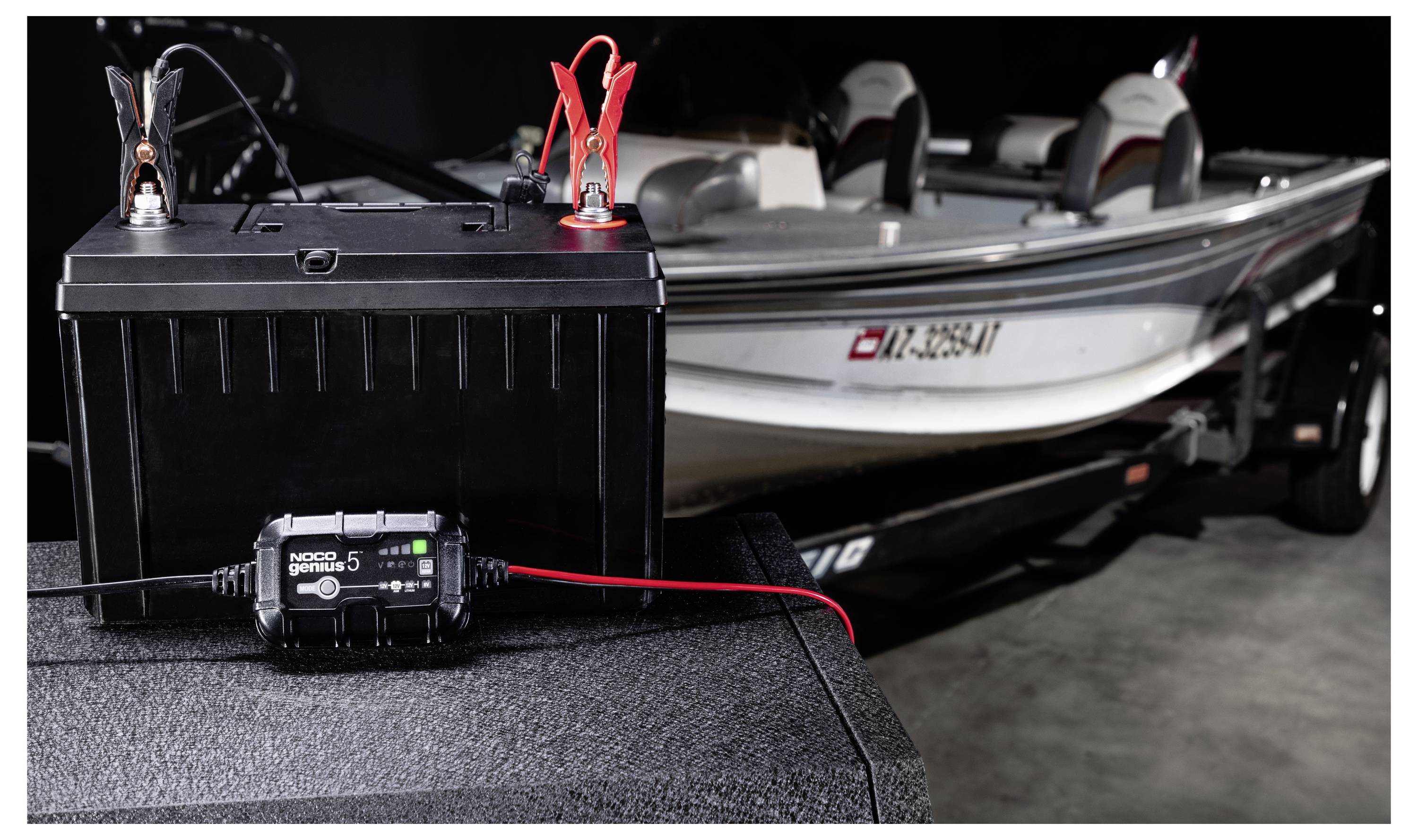 Tragbares Batterieladegerät mit roten und schwarzen Klemmen, die mit einer Bootbatterie verbunden sind und ein Fischerboot auf einem Anhänger mit Strom versorgen.
