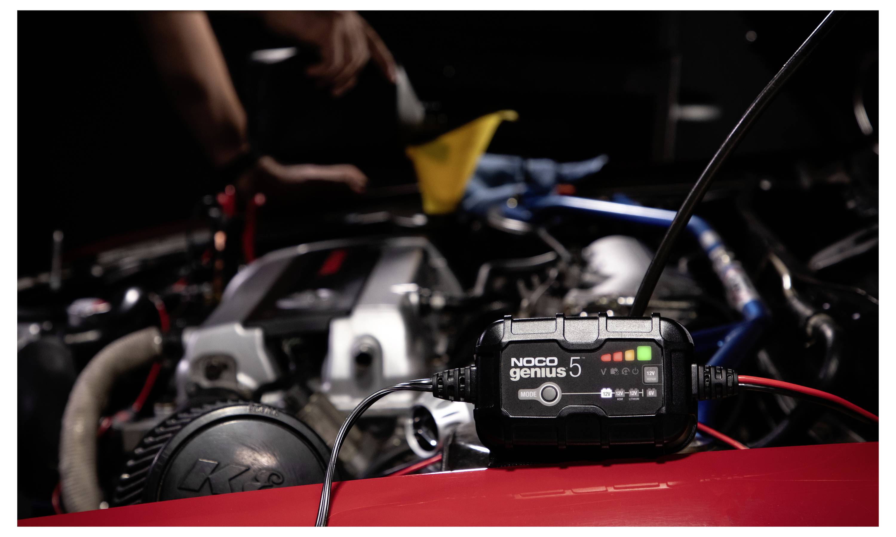 NOCO Genius 5 Batterieladegerät angeschlossen an einen Motorraum eines Autos, mit einer Person, die im Hintergrund arbeitet.