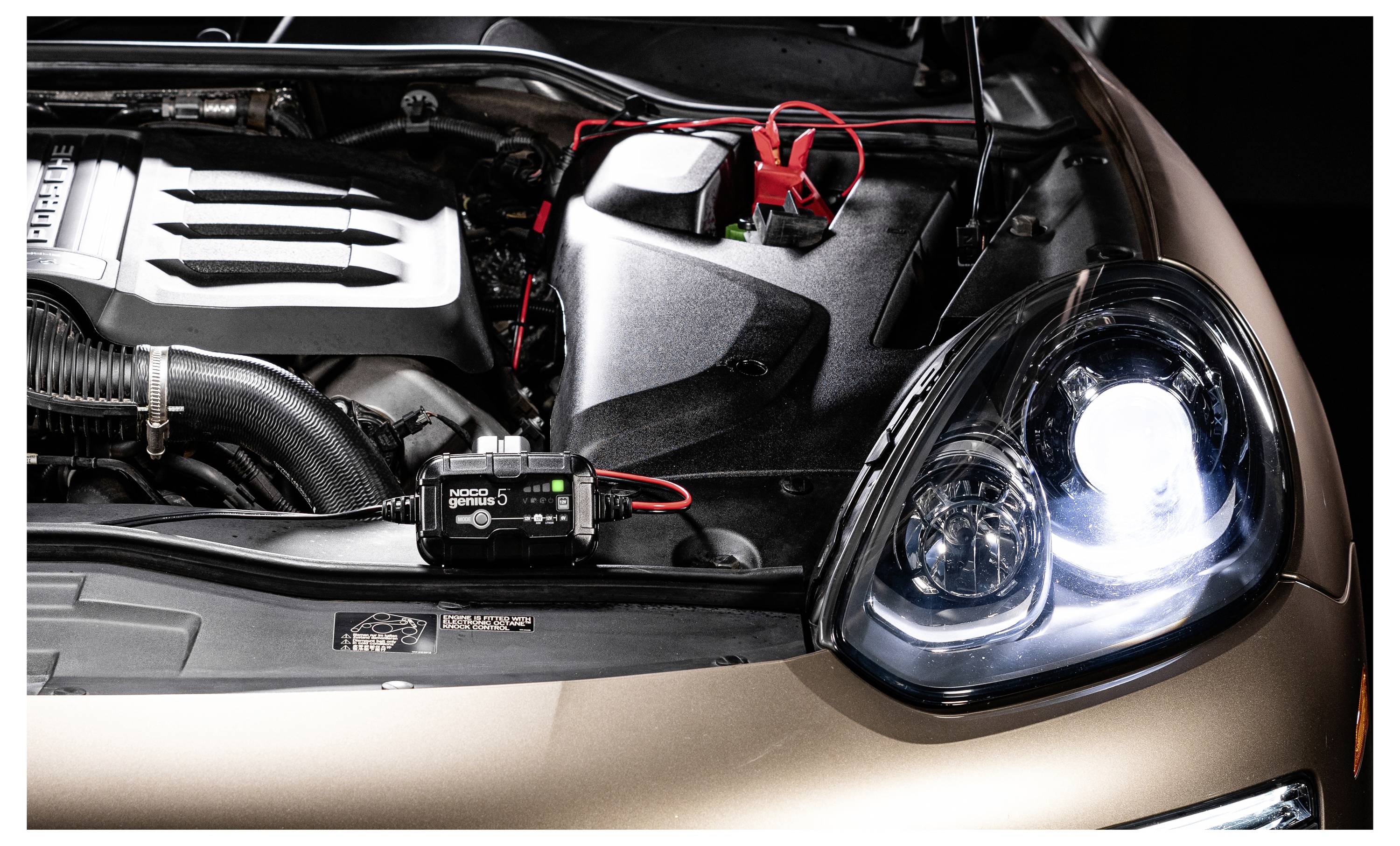 Motorraum eines Autos mit an Sprungstarter-/Batterieklemmkabeln angebrachten Kabeln in der Nähe des Scheinwerfers, was auf eine Starthilfe- oder Batterieladekonfiguration hindeutet.