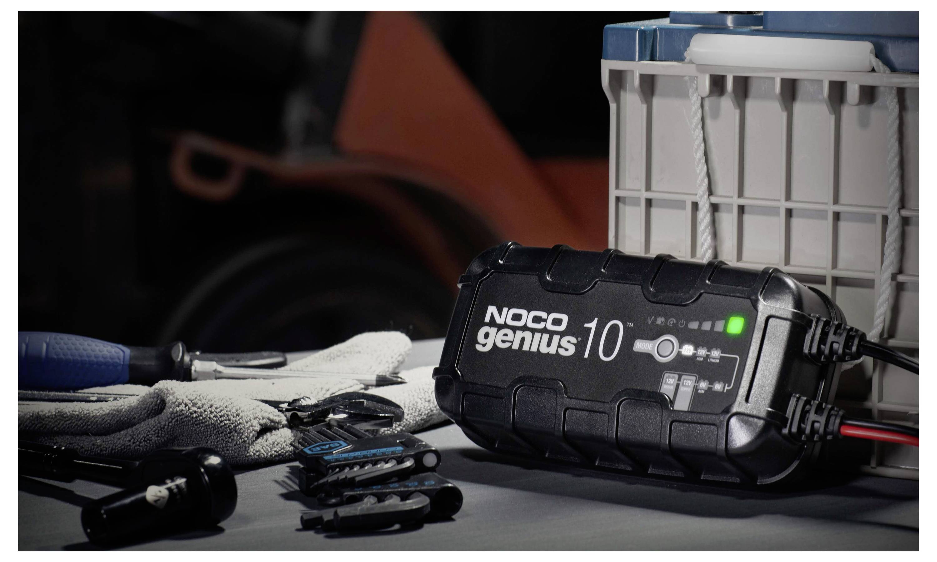 Ein NOCO Genius 10 Batterieladegerät sitzt neben Arbeitshandschuhen, Handwerkzeugen und einem Kunststoff-Aufbewahrungsbehälter auf einer Werkbank.