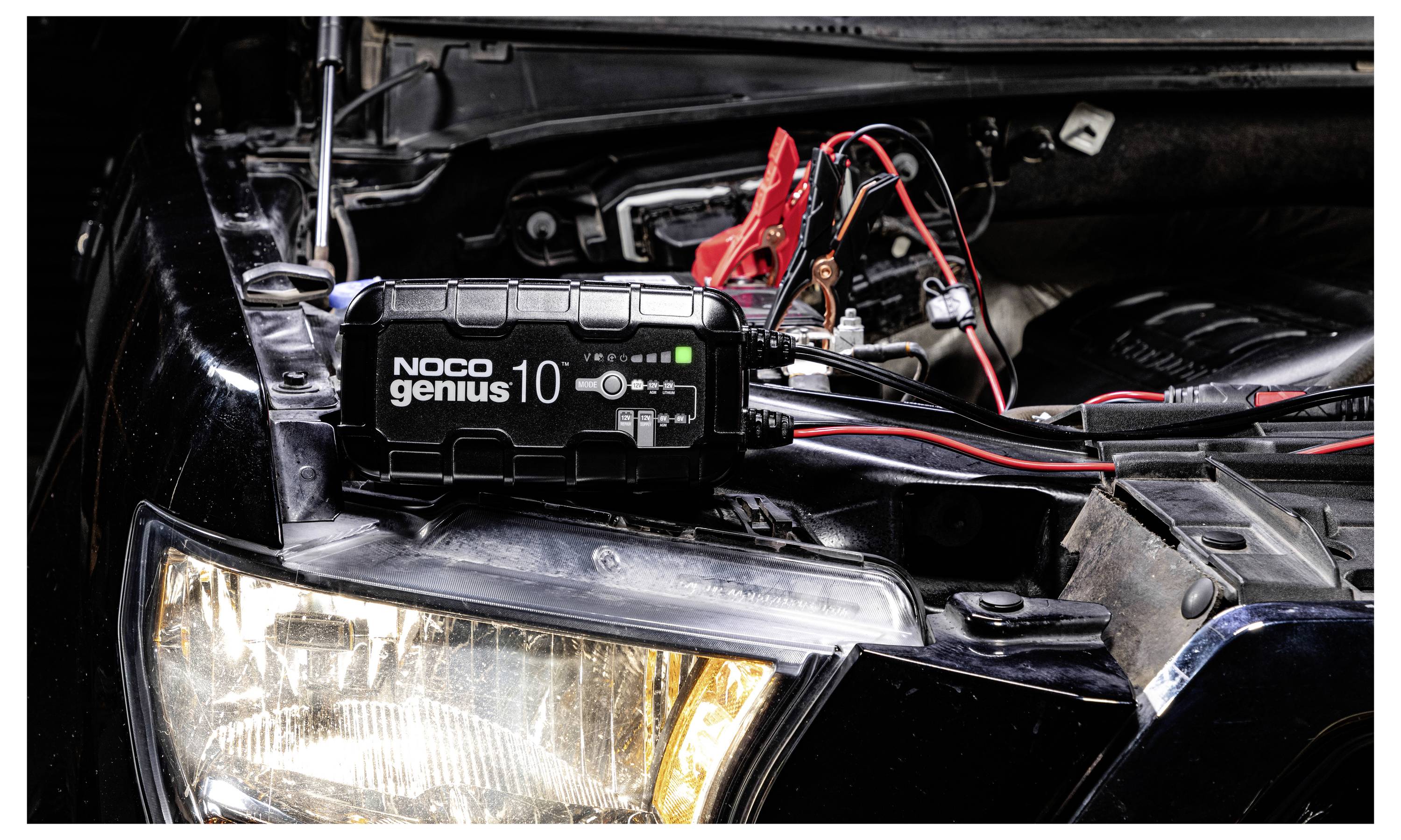 Tragbares NOCO Genius 10 Ladegerät, das mit einer Autobatterie in der offenen Motorhaube verbunden ist, mit roten und schwarzen Klemmen angebracht.