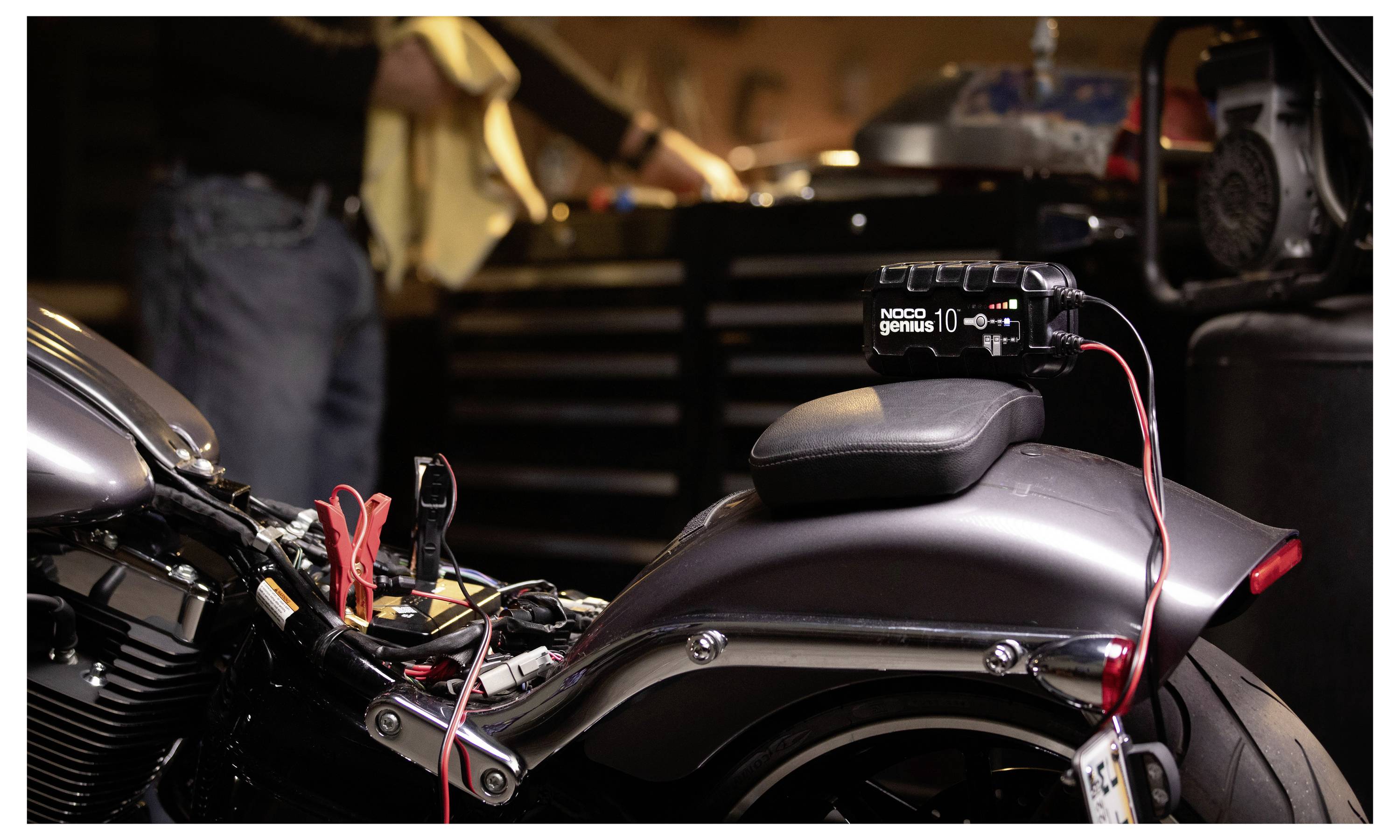 Motorradbatterieladegerät angeschlossen an einem geparkten Motorrad in einer Garage, mit roten und schwarzen Ladekabeln, die an die Batterie angeschlossen sind.
