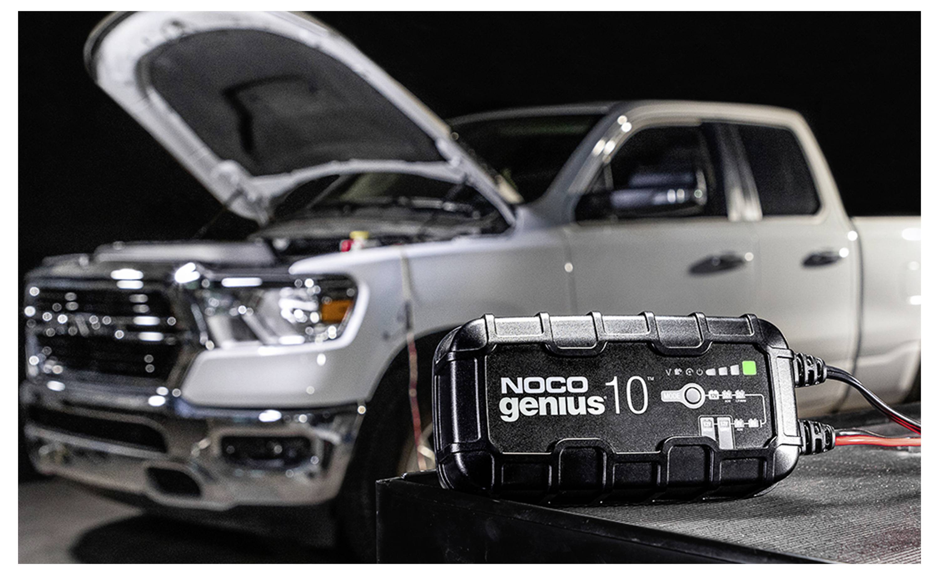 NOCO Genius10 Batterieladegerät im Vordergrund mit einem weißen Pickup-Truck dahinter und geöffneter Motorhaube, was auf das Laden oder die Wartung der Fahrzeugbatterie hindeutet.