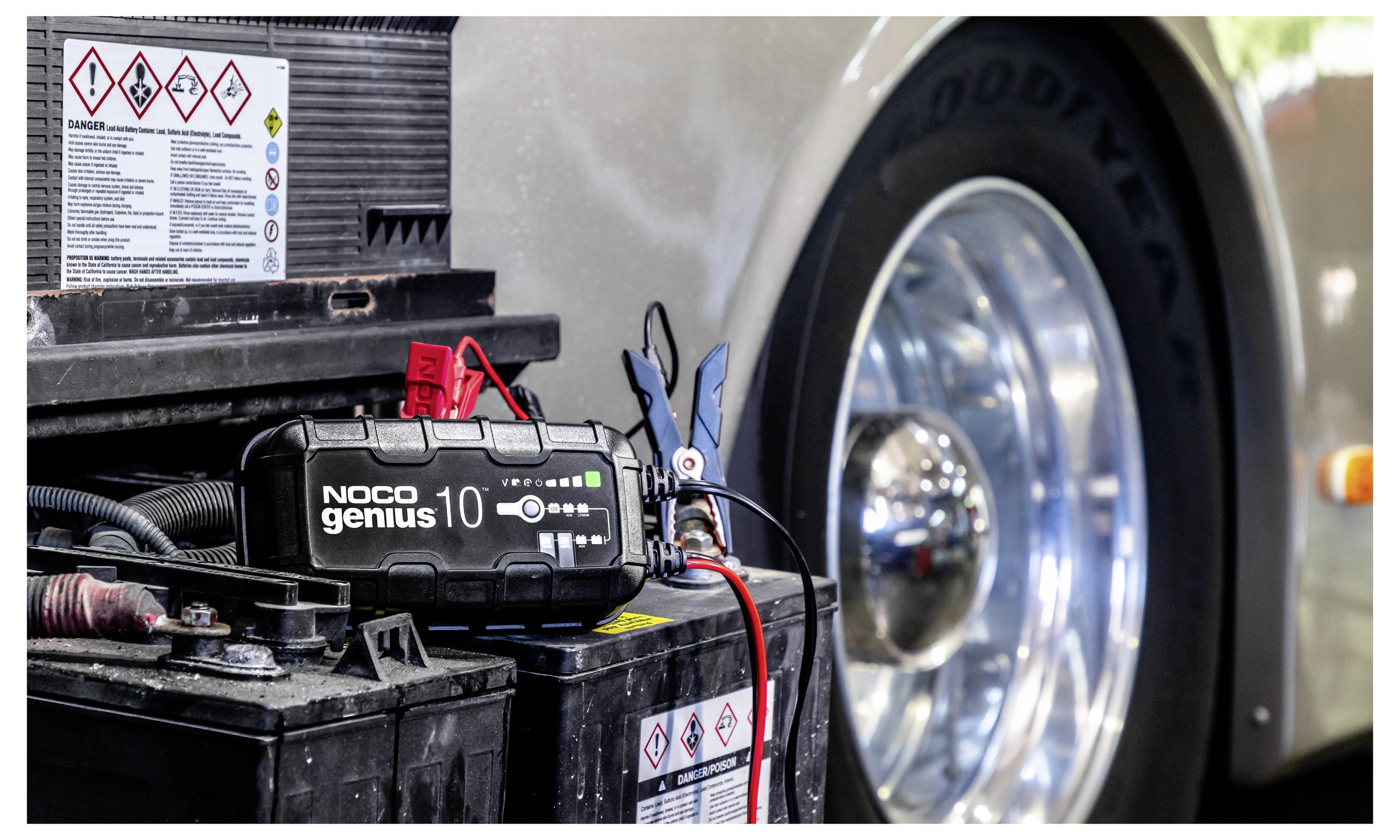 Tragbarer Starthilfebooster, der mit einer Autobatterie im Motorraum verbunden ist, wobei Starthilfekabel neben dem Vorderrad angebracht sind.