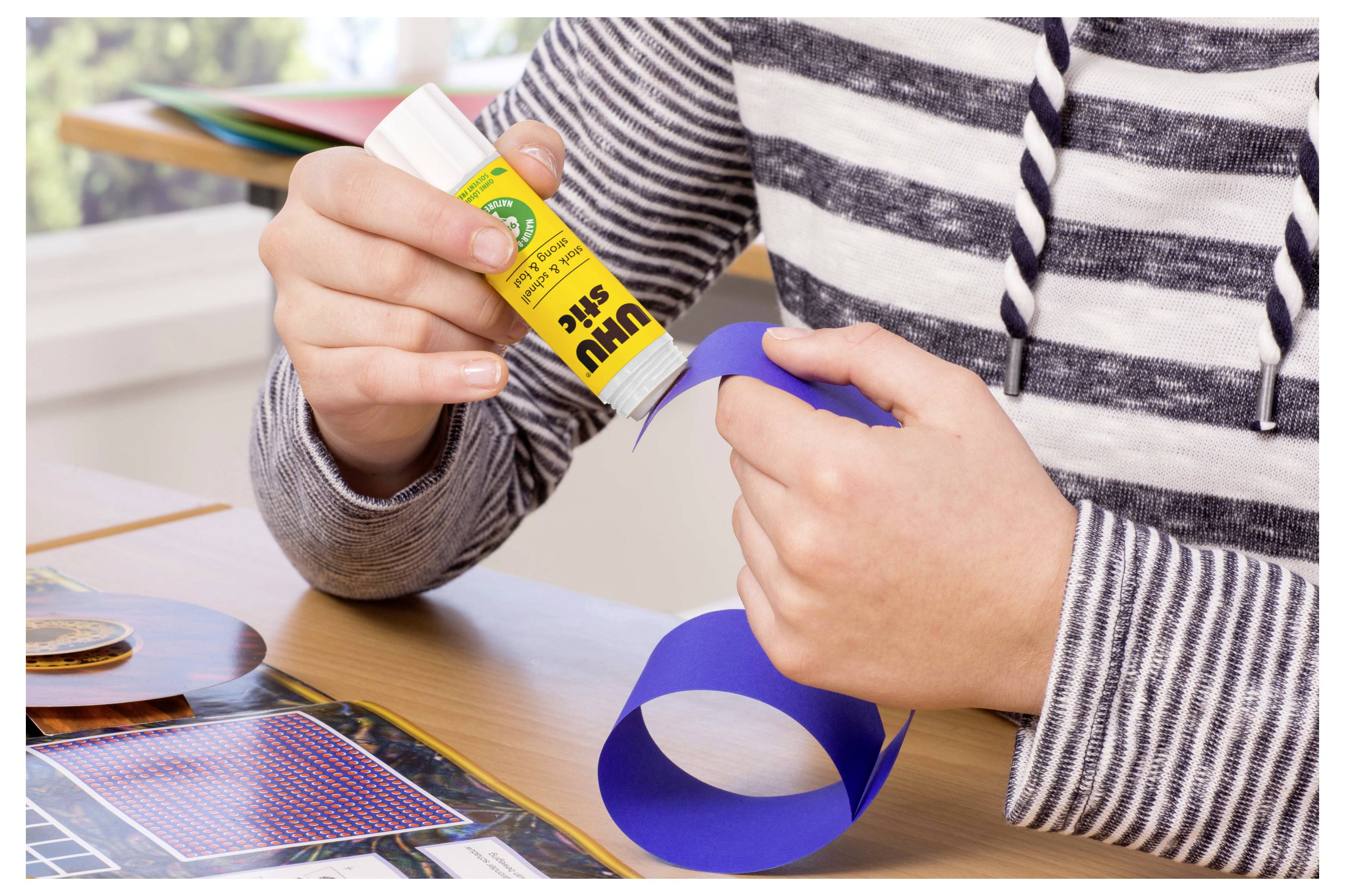 Eine Person trägt Klebstoff auf einen blauen Papierstreifen an einem Tisch auf und arbeitet an einem Bastelprojekt.
