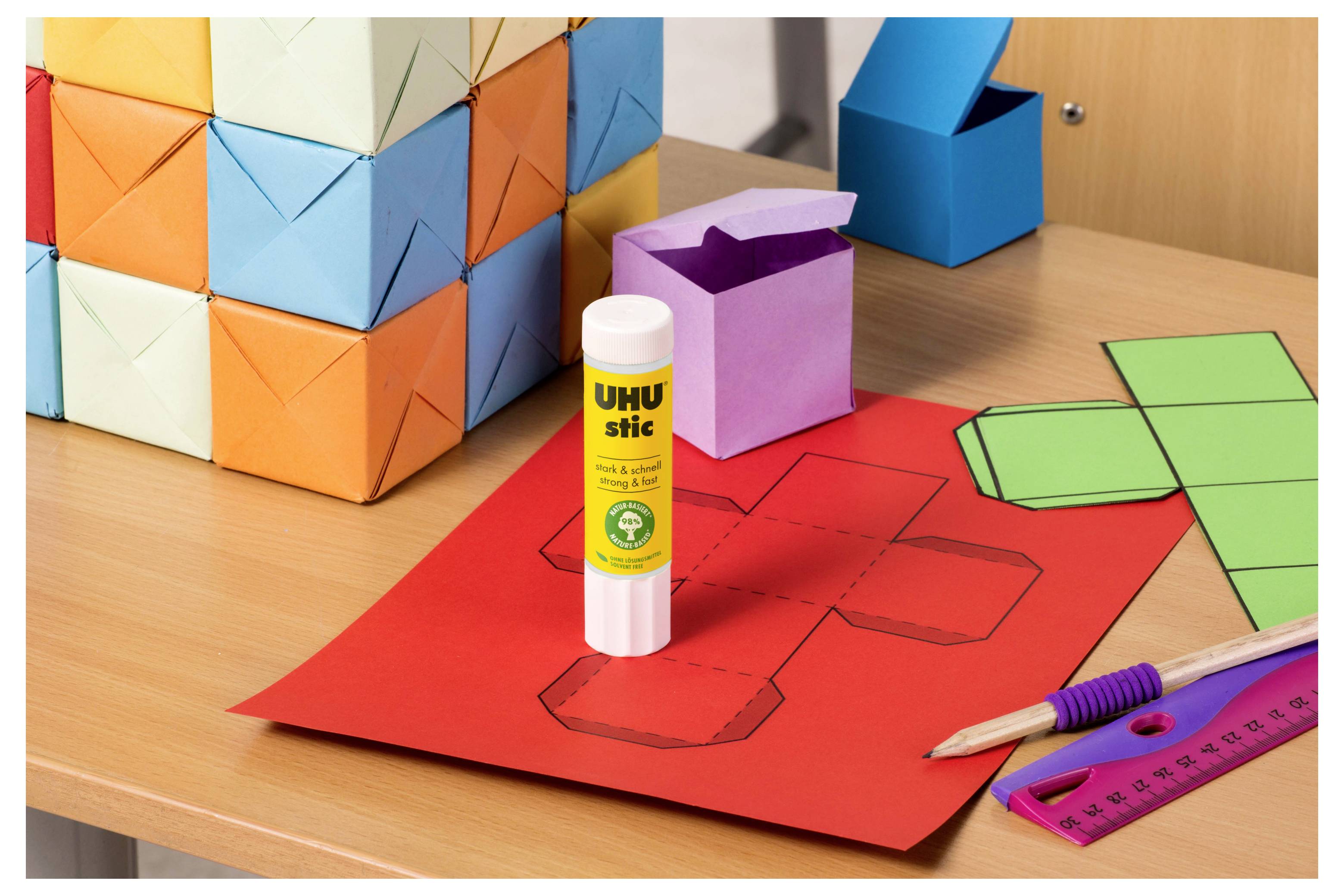 Bastelmaterialien auf einem Schreibtisch: ein Klebestift neben gefalteten Papierfiguren, farbige Papiervorlagen, ein Bleistift, ein Lineal und ein Origami-Würfel im Hintergrund.