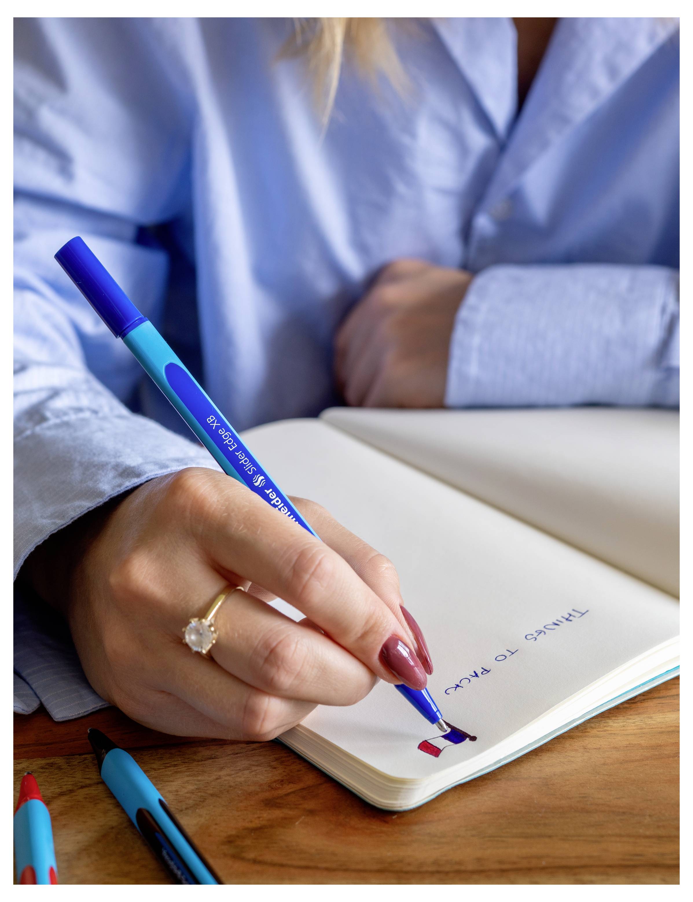 Eine Person schreibt in einem Notizbuch mit einem blauen Stift, während sie an einem Holzschreibtisch sitzt, mit einer offenen Seite und weiteren Stiften in der Nähe.