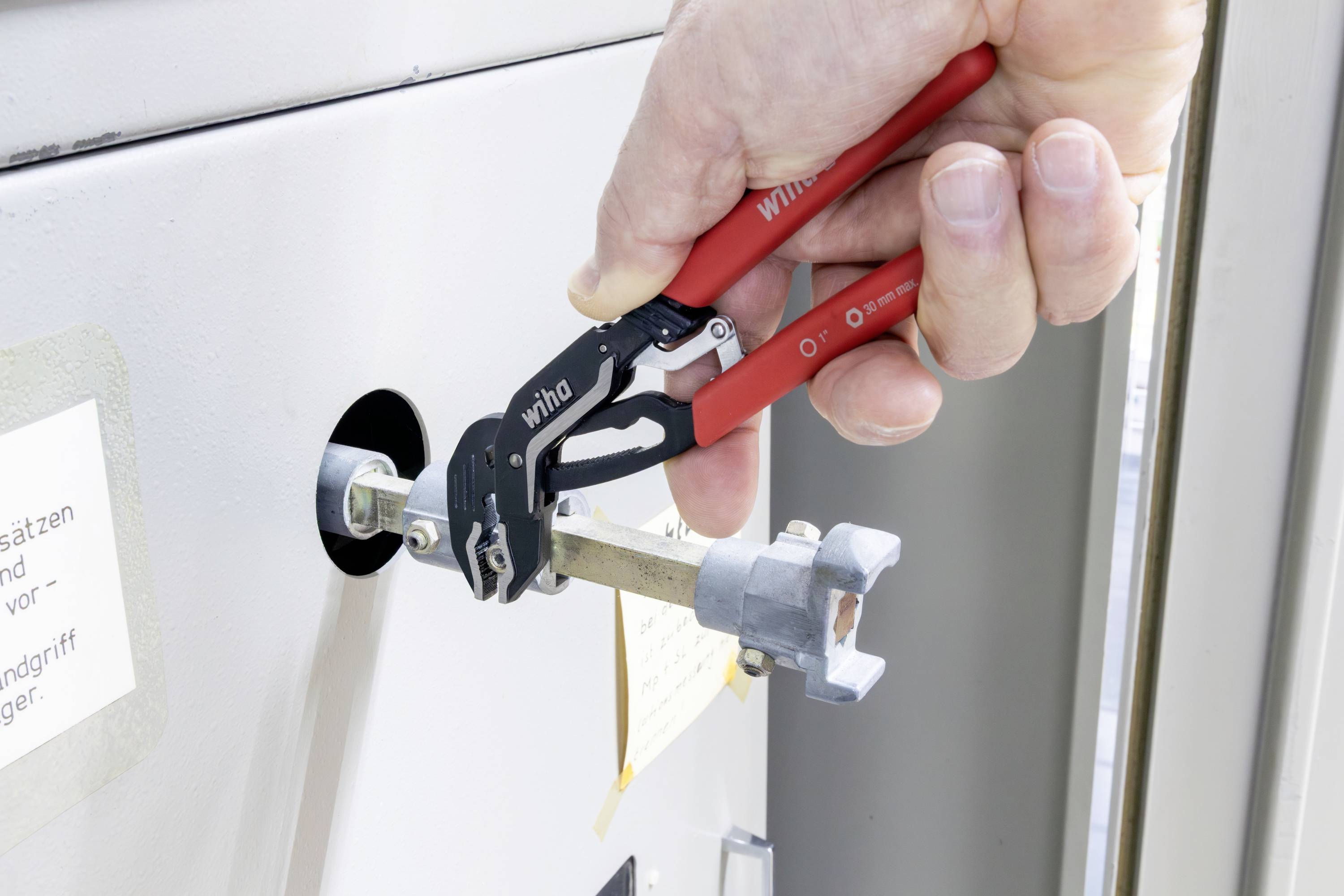 Hand mit roten Zangen, die ein Metallventil oder Schloss an einem weißen Schrank oder Bedienfeld dreht.