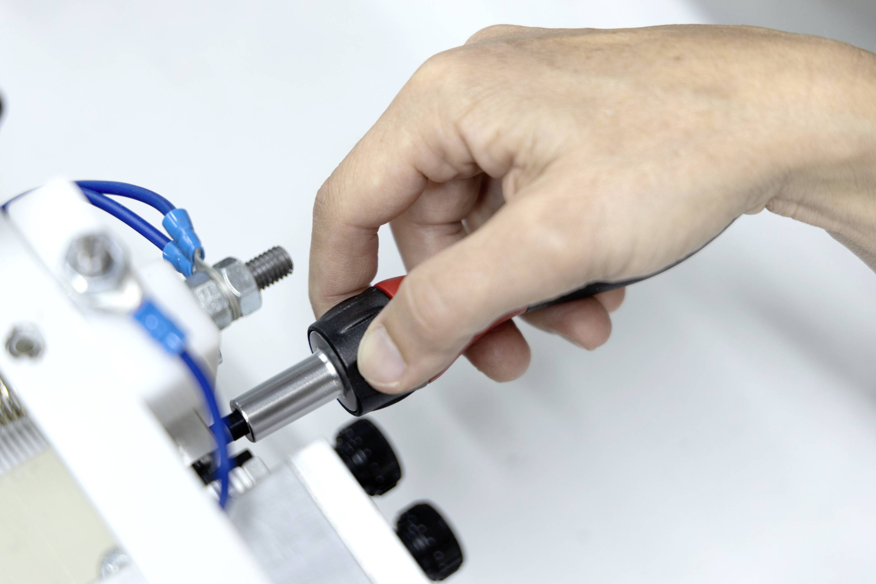 Eine Hand führt einen schwarzen Stecker in eine weiße Anschlussöffnung einer Maschine ein, neben blauen Kabeln und einer Metallarmatur.