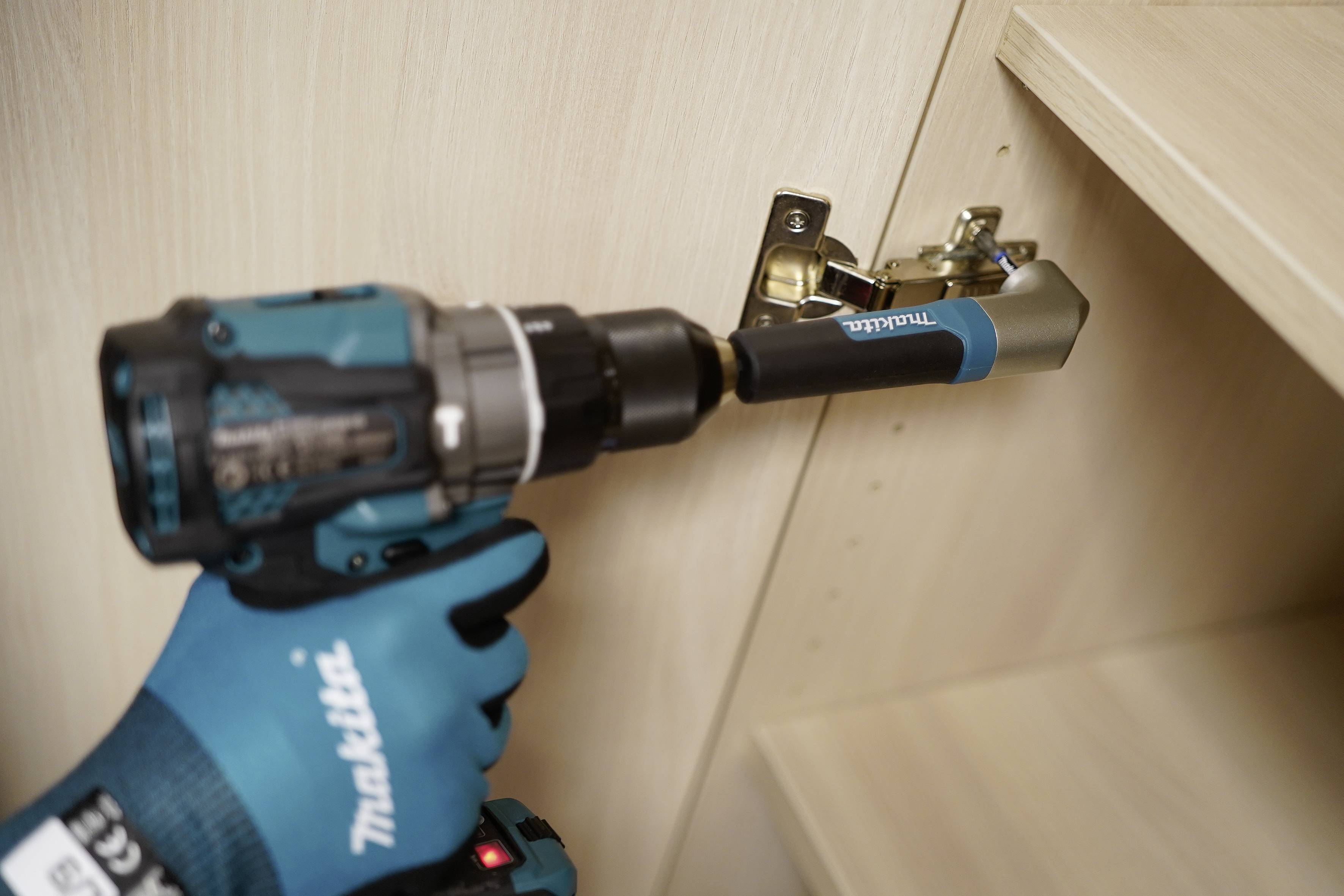 Eine behandschuhte Hand verwendet einen Makita-Akku-Bohrer, um ein Schrankschanier in einem Holzschrank einzubauen.