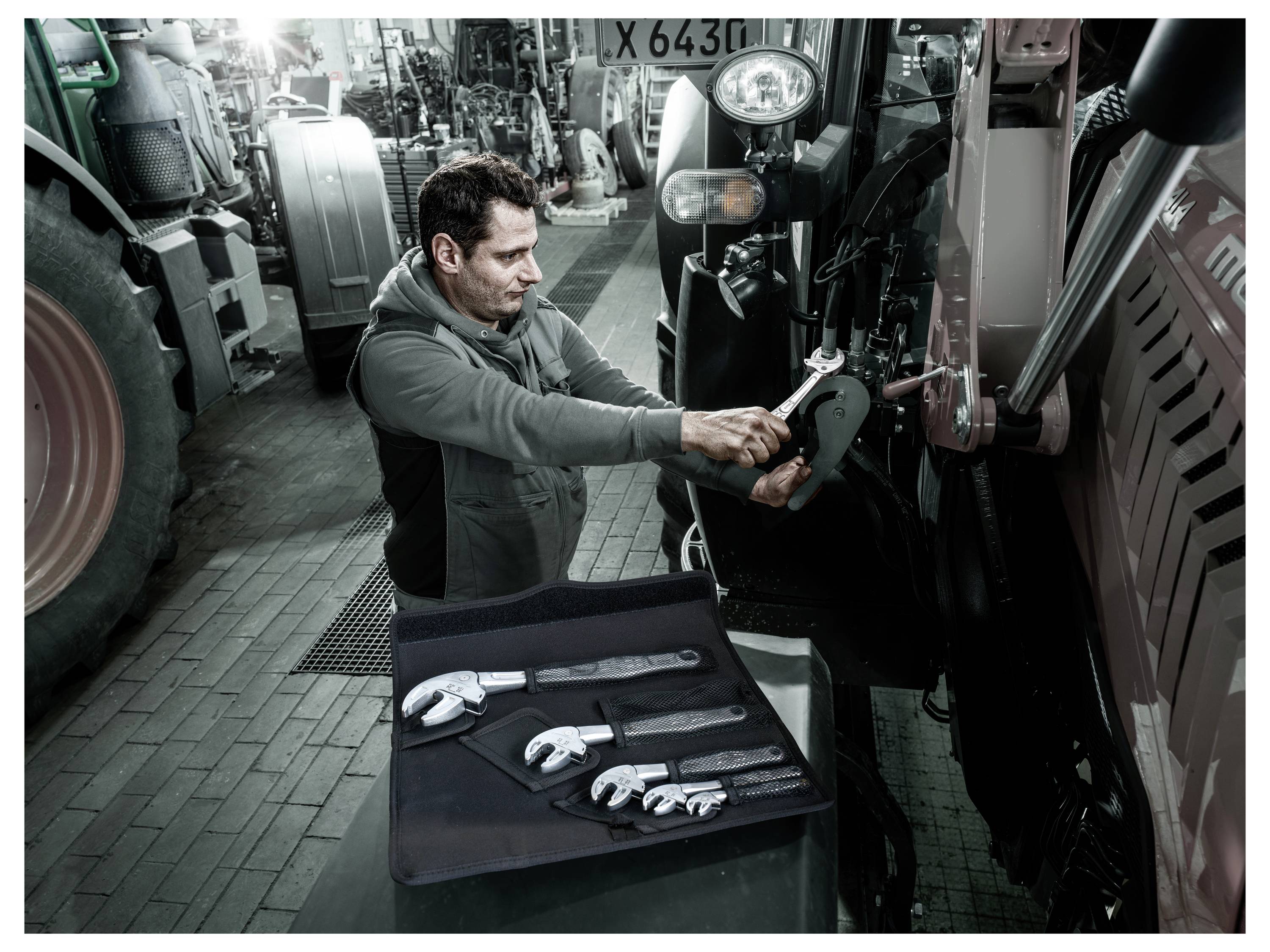 Mechaniker, der einen Schraubenschlüssel verwendet, um einen Traktor in einer Werkstatt zu warten, mit weiteren Werkzeugen auf einem nahegelegenen Wagen angeordnet.