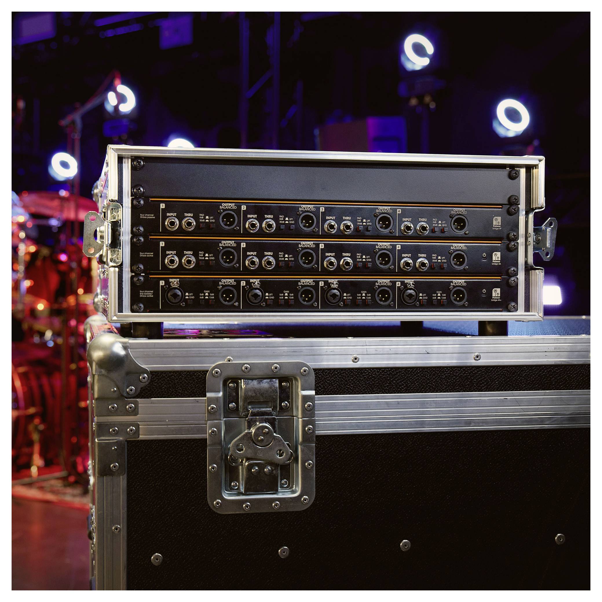 Auf einem schwarzen Flightcase gestapelte Rack-Audigeräte in einer Bühnen- oder Konzertumgebung, mit unscharfen Schlagzeugen und farbigen Lichtern im Hintergrund.