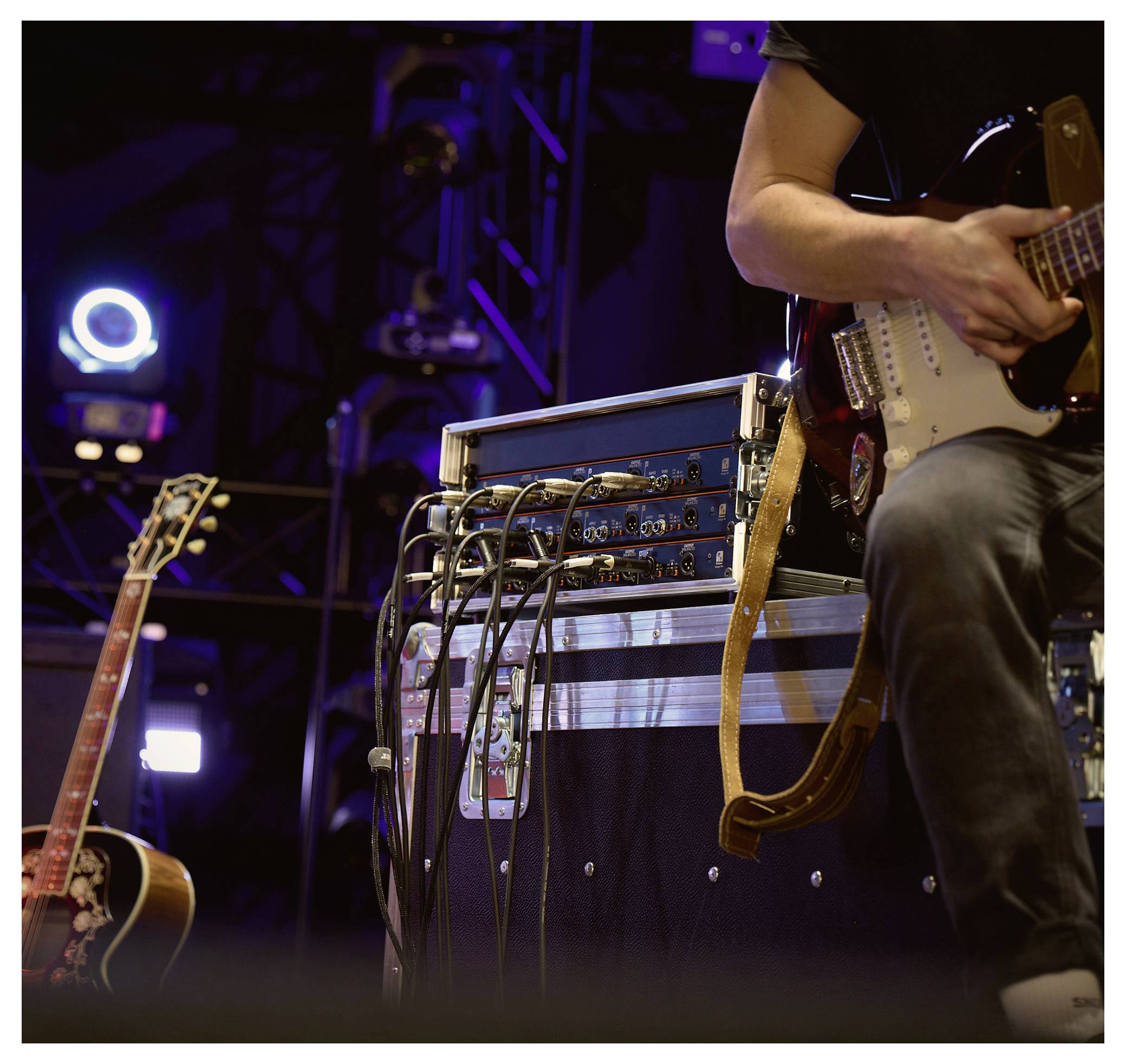 Bühnenmusikeaufbau mit einem Gitarristen, der eine E-Gitarre neben einem Geräteständer und Kabeln spielt, mit einer Akustikgitarre im Hintergrund.