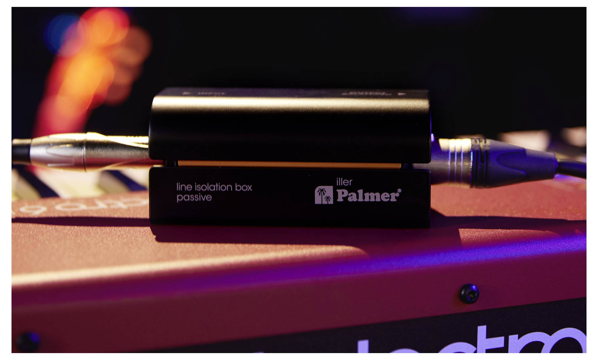 Nahaufnahme einer Palmer Passive Line Isolation Box mit Audiokabeln, die an beiden Enden angeschlossen sind, auf einer roten Oberfläche unter Bühnenleuchtung platziert.