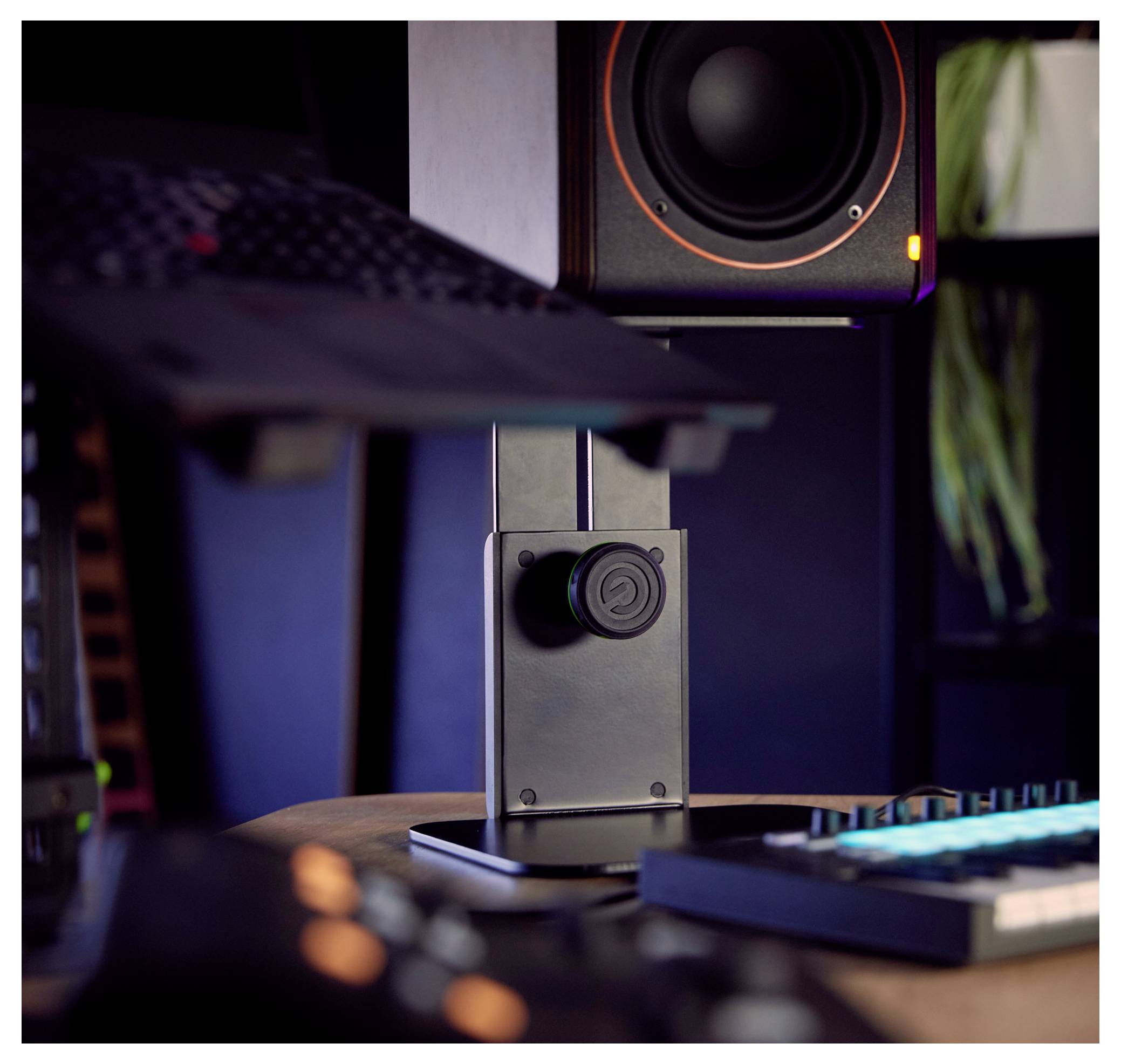 Nahaufnahme eines schwarzen Desktop-Lautsprecherständers mit einem Audio-Regler neben einer Tastatur und einem Monitor in einer schwach beleuchteten Studio-Einrichtung.