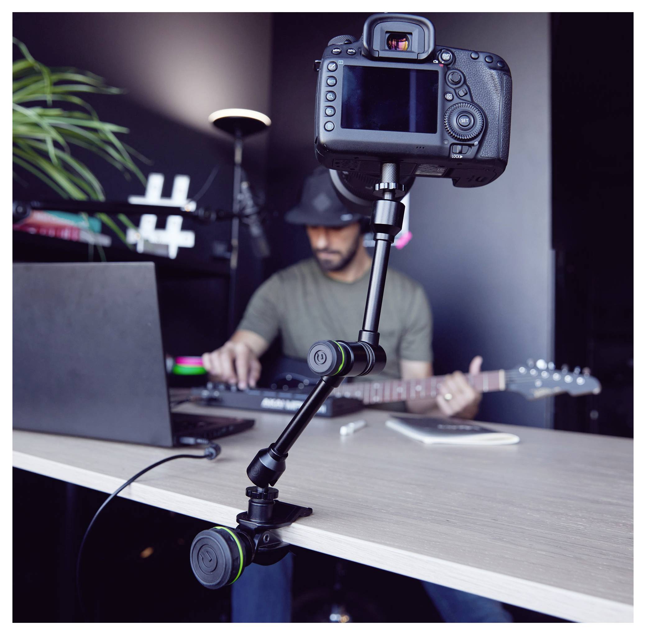 Kamera auf einem Stativ nimmt einen Mann auf, der Musik auf einem Laptop bearbeitet, während er eine Gitarre in der Hand hält, in einem Studioarbeitsplatz.