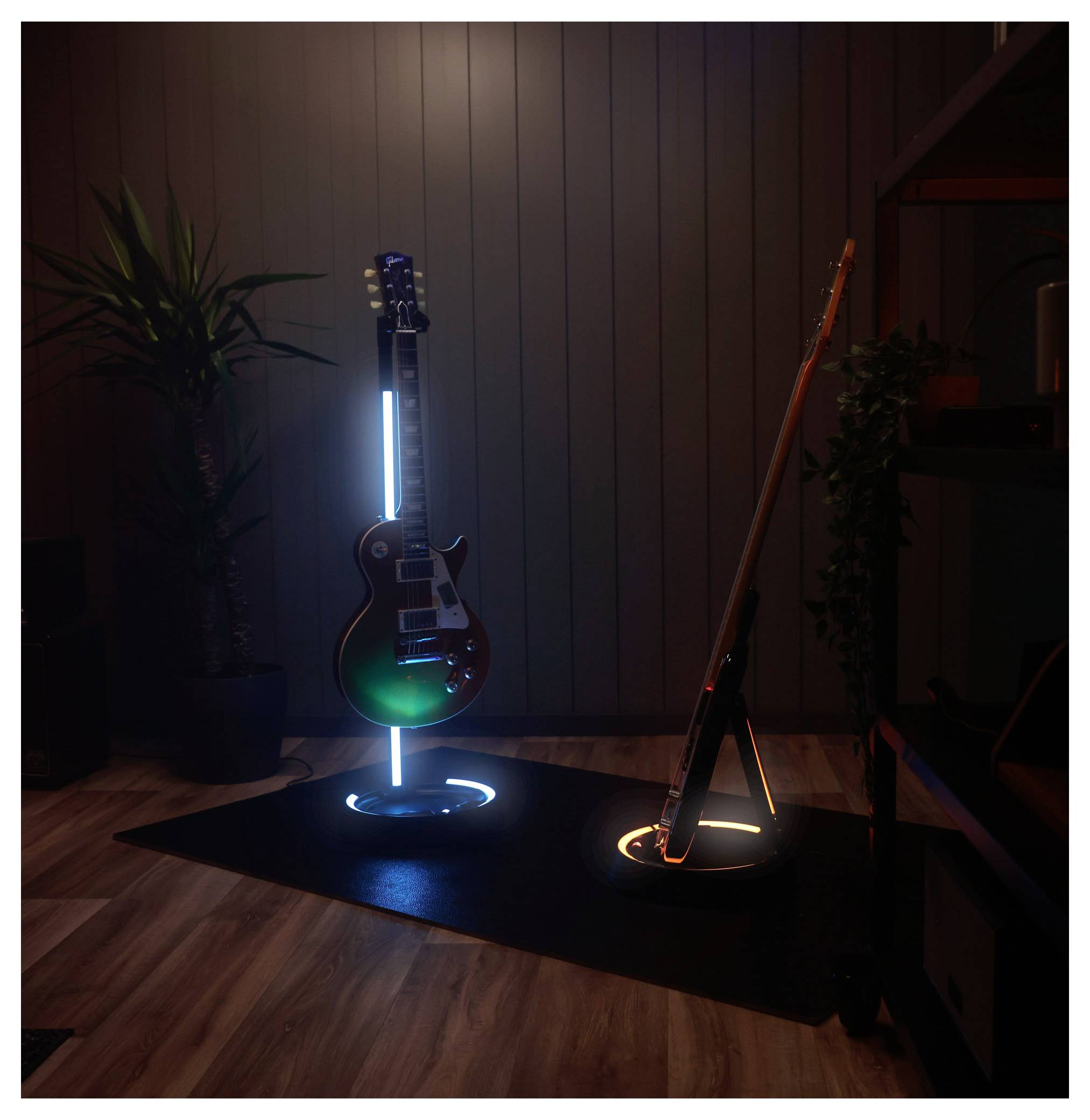 Zwei E-Gitarren auf beleuchteten Bodenständern in einem dunklen Raum, eine blau beleuchtet und eine orange, mit Pflanzen und Holzverkleidung im Hintergrund.