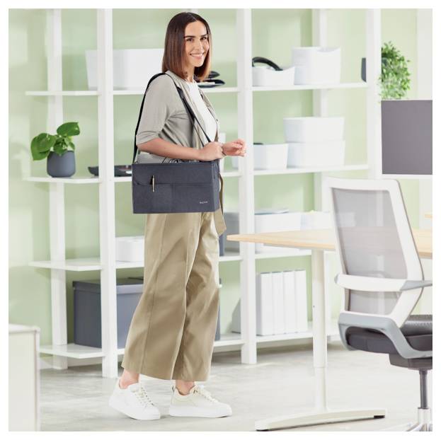 Frau, die in einem hellen Büro steht und eine dunkle Umhängetasche hält, mit weißen Regalen und Schreibtischen im Hintergrund.