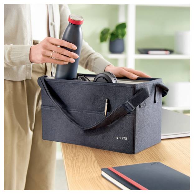 Eine Person packt eine Wasserflasche in eine graue Leitz-Tragetasche auf einem Schreibtisch, mit Kopfhörern darin und einem Notizbuch in der Nähe.