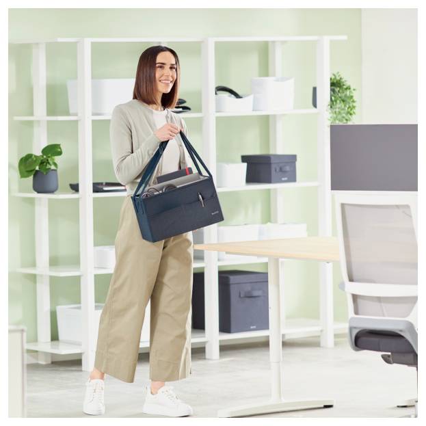 Frau, die in einem modernen Büro steht, einen schwarzen Aktenkoffer hält, lächelt neben einem Schreibtisch und einem Regal.