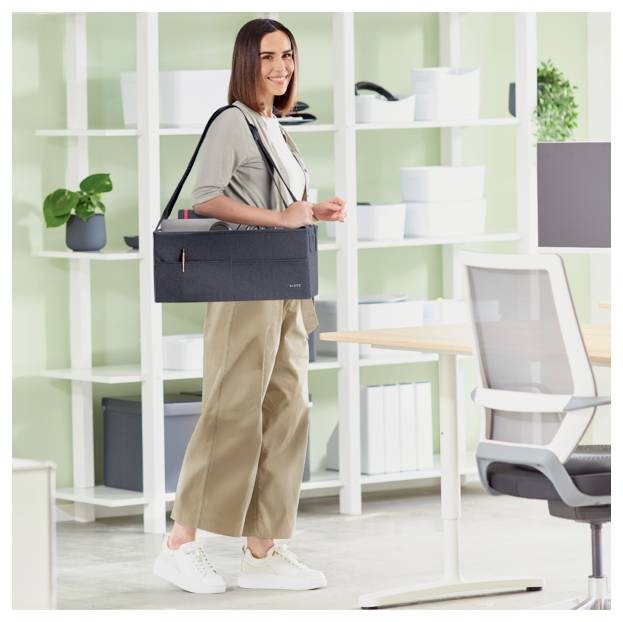 Frau, die in einem hellen Büro steht und eine schwarze Umhängetasche hält, neben einem Schreibtisch und einem Regal.