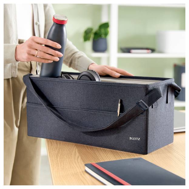 Eine Person legt eine wiederverwendbare Flasche in einen dunklen Stoff-Aufbewahrungsbehälter auf einem Schreibtisch, mit Kopfhörern darin und einem Notizbuch in der Nähe.