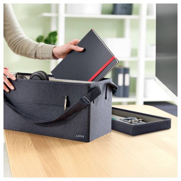 Eine Person legt ein schwarzes Notizbuch in eine graue Stofflagerkiste auf einem Schreibtisch, mit Kopfhörern und kleinen Bürobedarfsartikeln daneben.