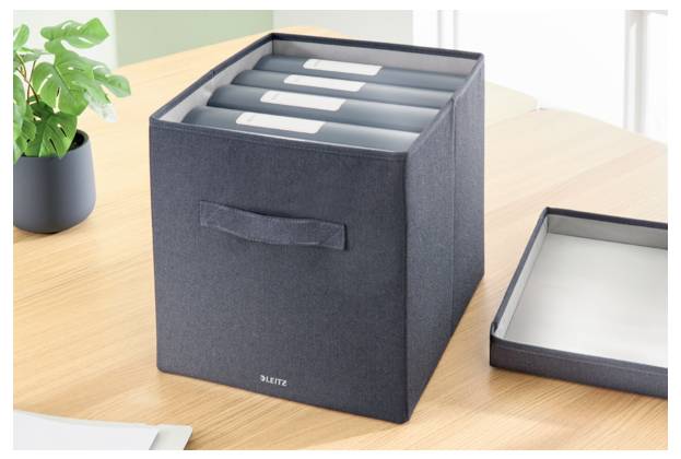 Aufbewahrungsbox aus dunklem Stoff mit Griffen und beschrifteten Ordnern auf einem Holzschreibtisch neben einer Pflanze und einem Deckel.