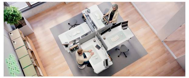 Draufsicht auf zwei Personen, die an benachbarten Schreibtischen in einem modernen offenen Arbeitsbereich mit Computermonitoren und Stühlen arbeiten.