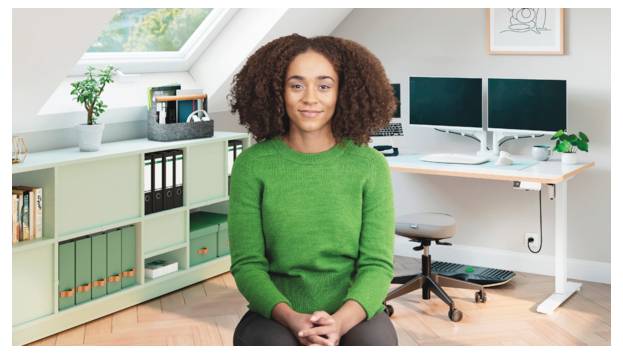 Frau in einem Homeoffice, sitzend und lächelnd vor zwei Monitoren, Regalen und Pflanzen.