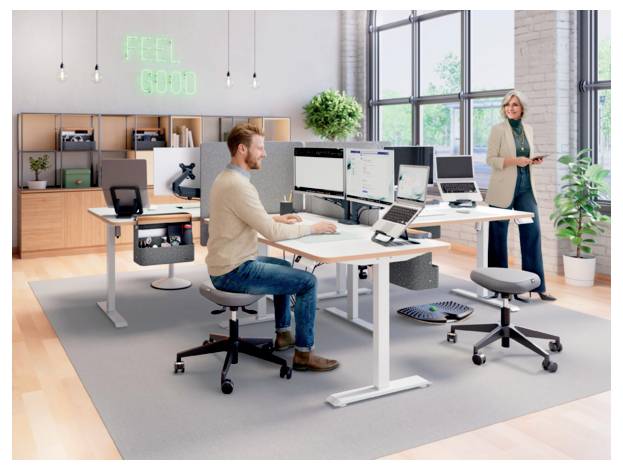 Modernes Großraumbüro mit einem Mann, der an einem Stehschreibtisch mit mehreren Monitoren arbeitet, während eine Frau in der Nähe steht; ein Neonschild liest „FEEL GOOD