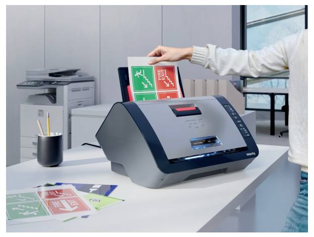 Eine Person führt ein Blatt gedruckter Etiketten in einen Desktop-Etikettendrucker auf einem Bürotisch ein, mit einem Kopierer und Etikettenmuster in der Nähe.