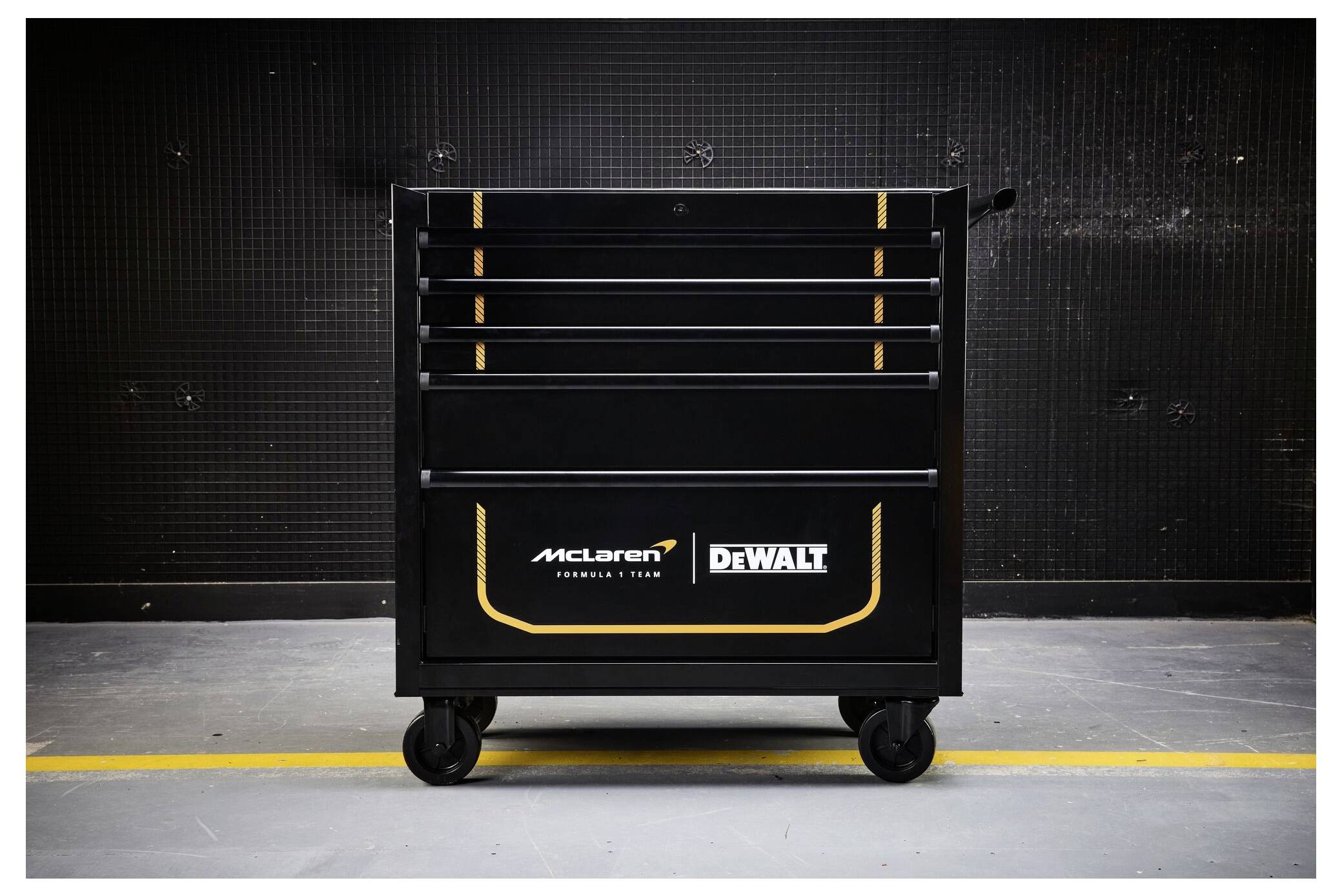 Schwarze fahrbarer Werkzeugkasten mit McLaren- und DEWALT-Logos, zentriert gegen eine dunkle Industriewand.