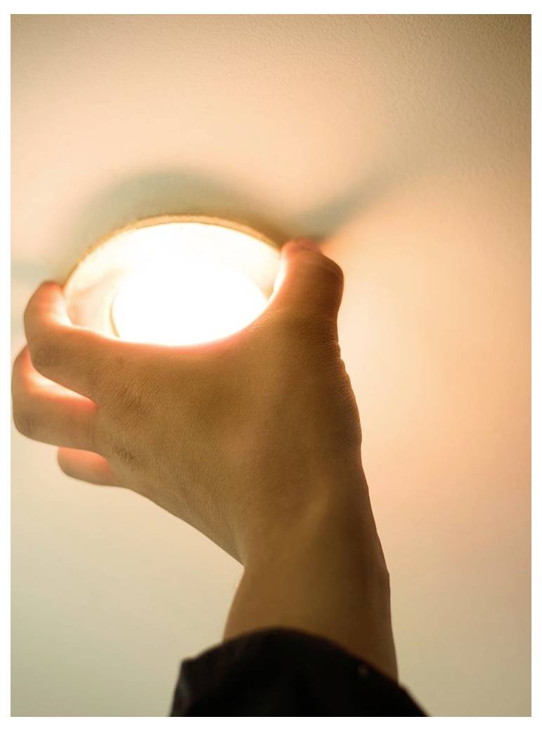Eine Hand hält eine leuchtende Glühbirne nah an die Wand, wodurch in einem dunklen Raum ein heller warmer Lichthof entsteht.