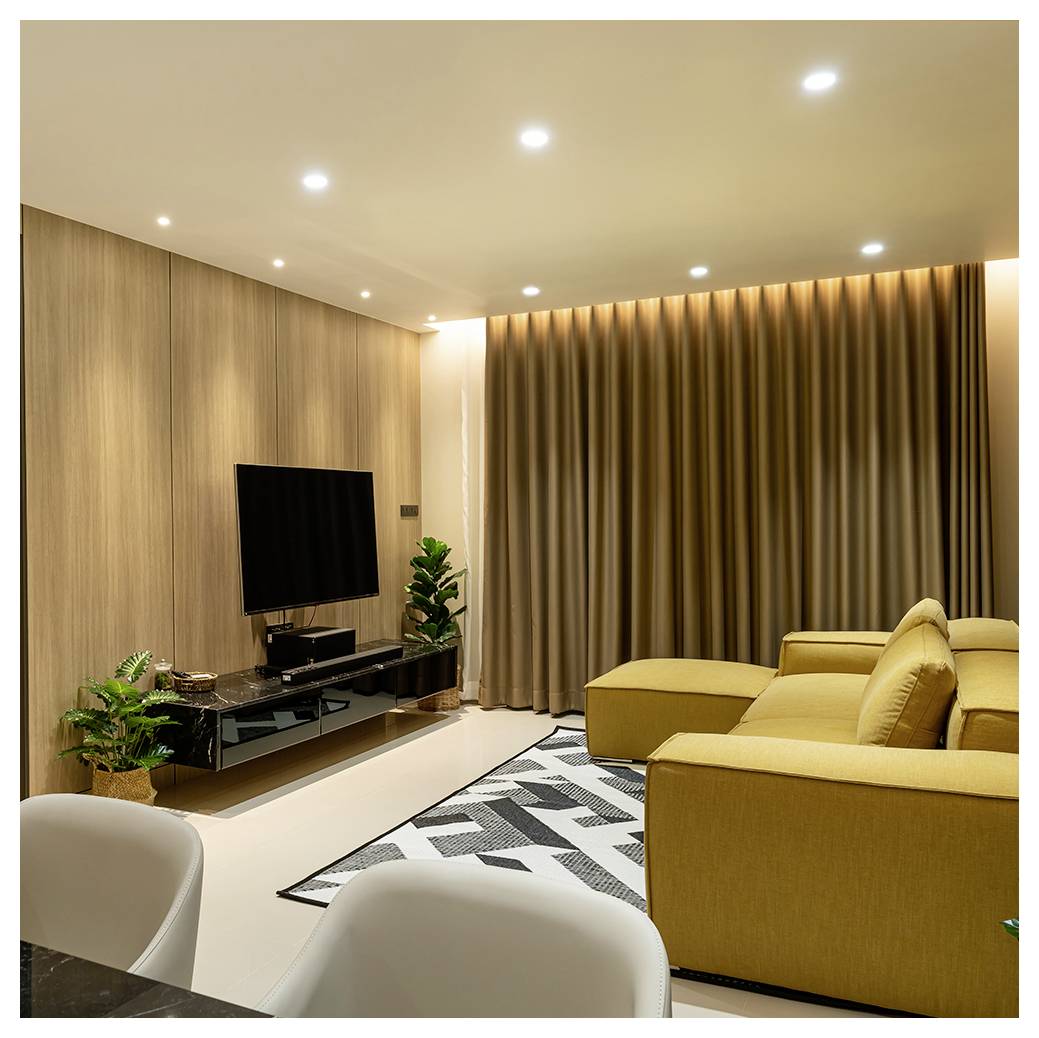 Modernes Wohnzimmer mit gelbem Ecksofa, an der Wand montiertem Fernseher, Holzvertäfelung, beigen Gardinen, Pflanzen und gemustertem Teppich.